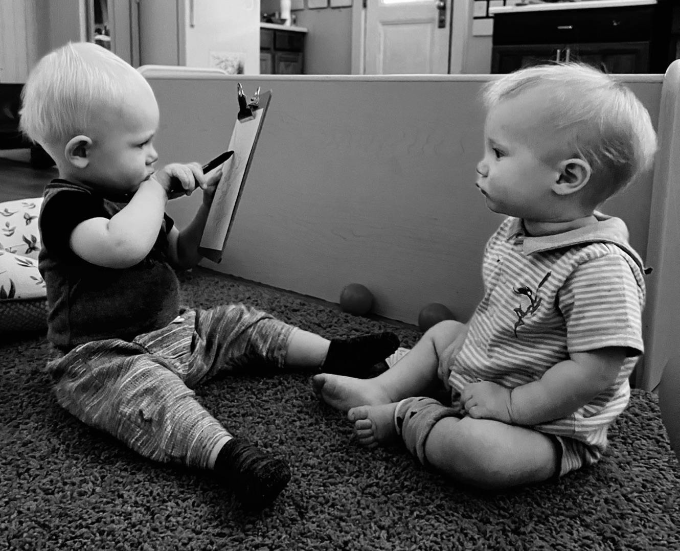 Two children sitting on a carpet, one holding a marker to a clipboard