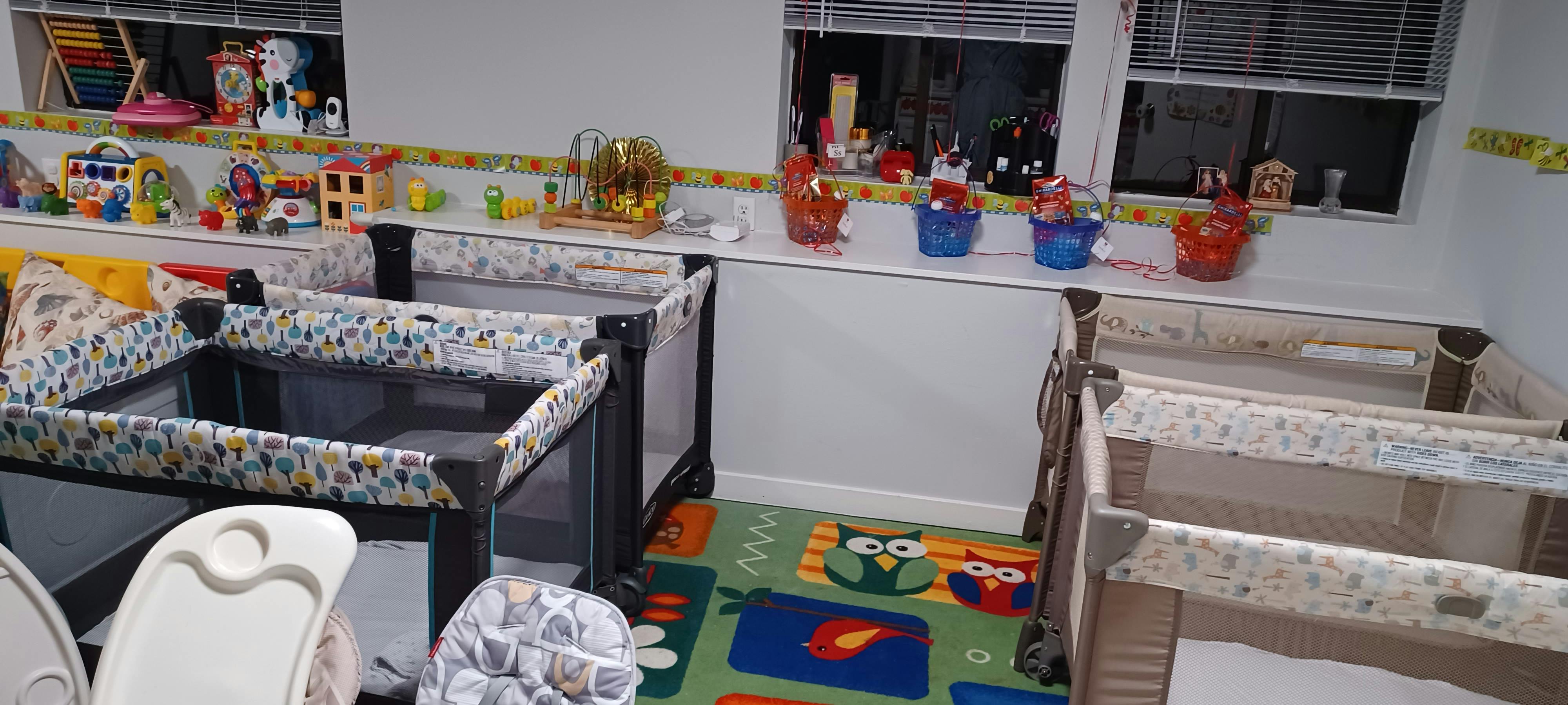 Multiple playpens in a room with a colorful rug and toys on a shelf