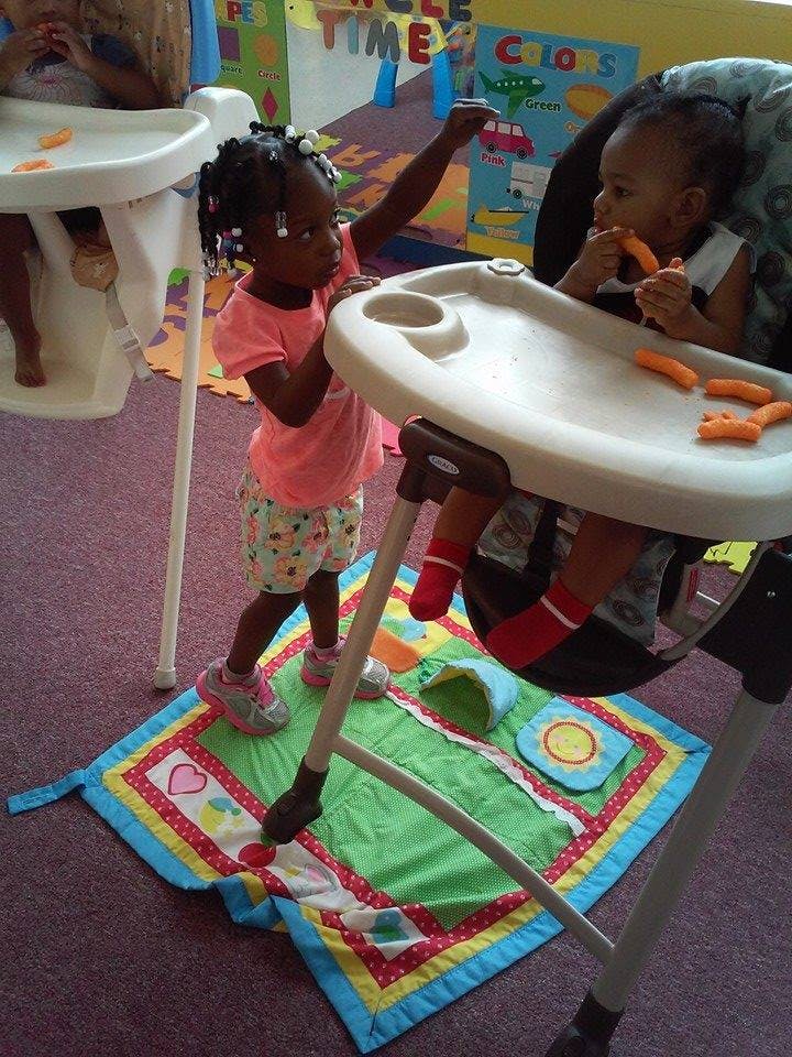 Child standing next to a high chair with another child eating in it