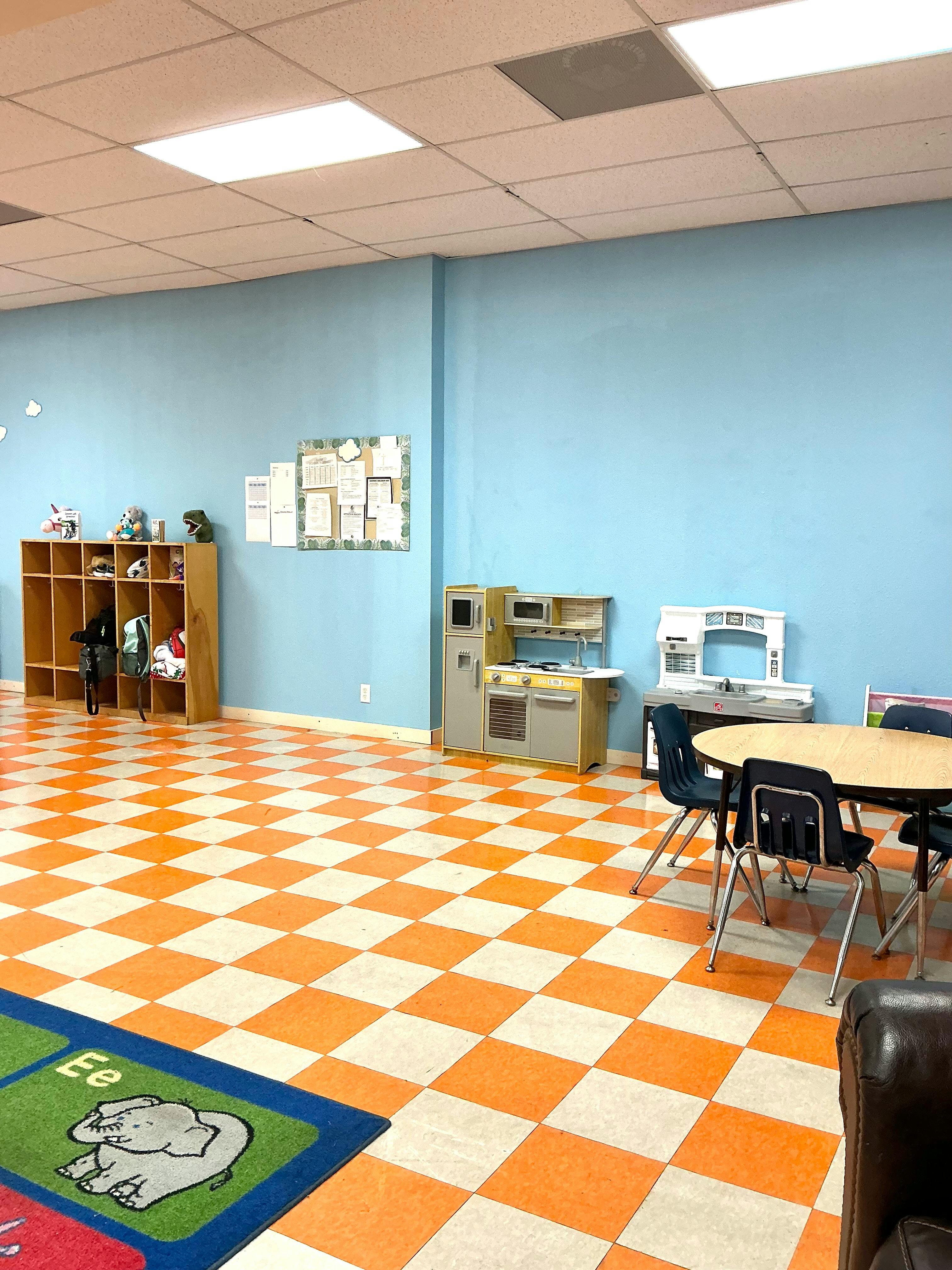 Empty classroom with checkered floor, cubbies, play kitchen, and small tables