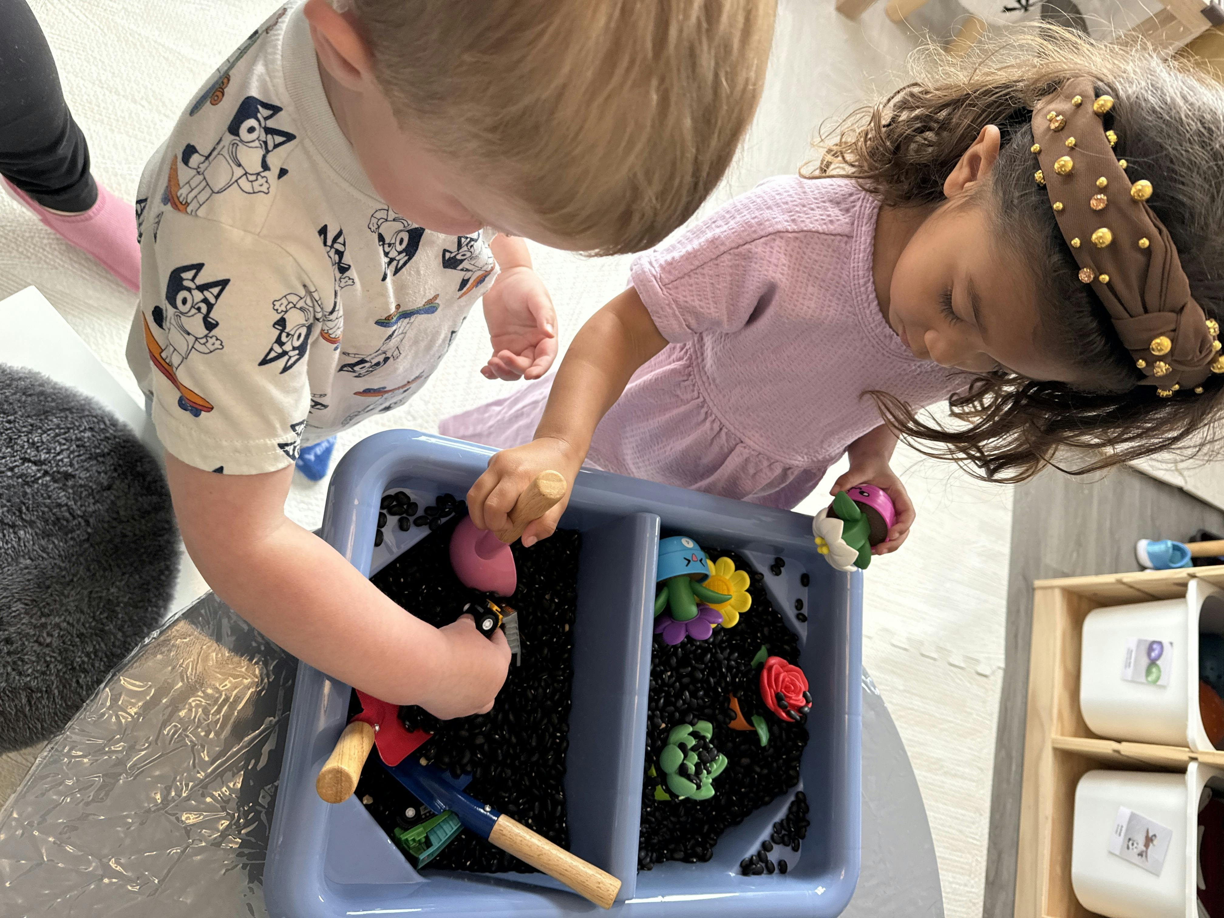 Two children playing with toys in a sensory bin filled with black beans