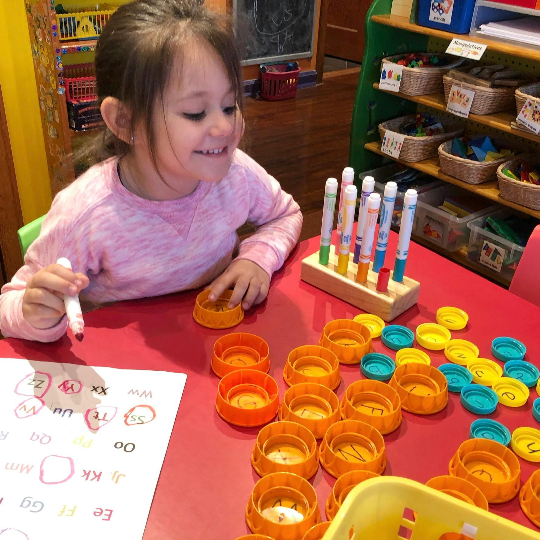 Child writing on paper with a marker, surrounded by colorful bottle caps