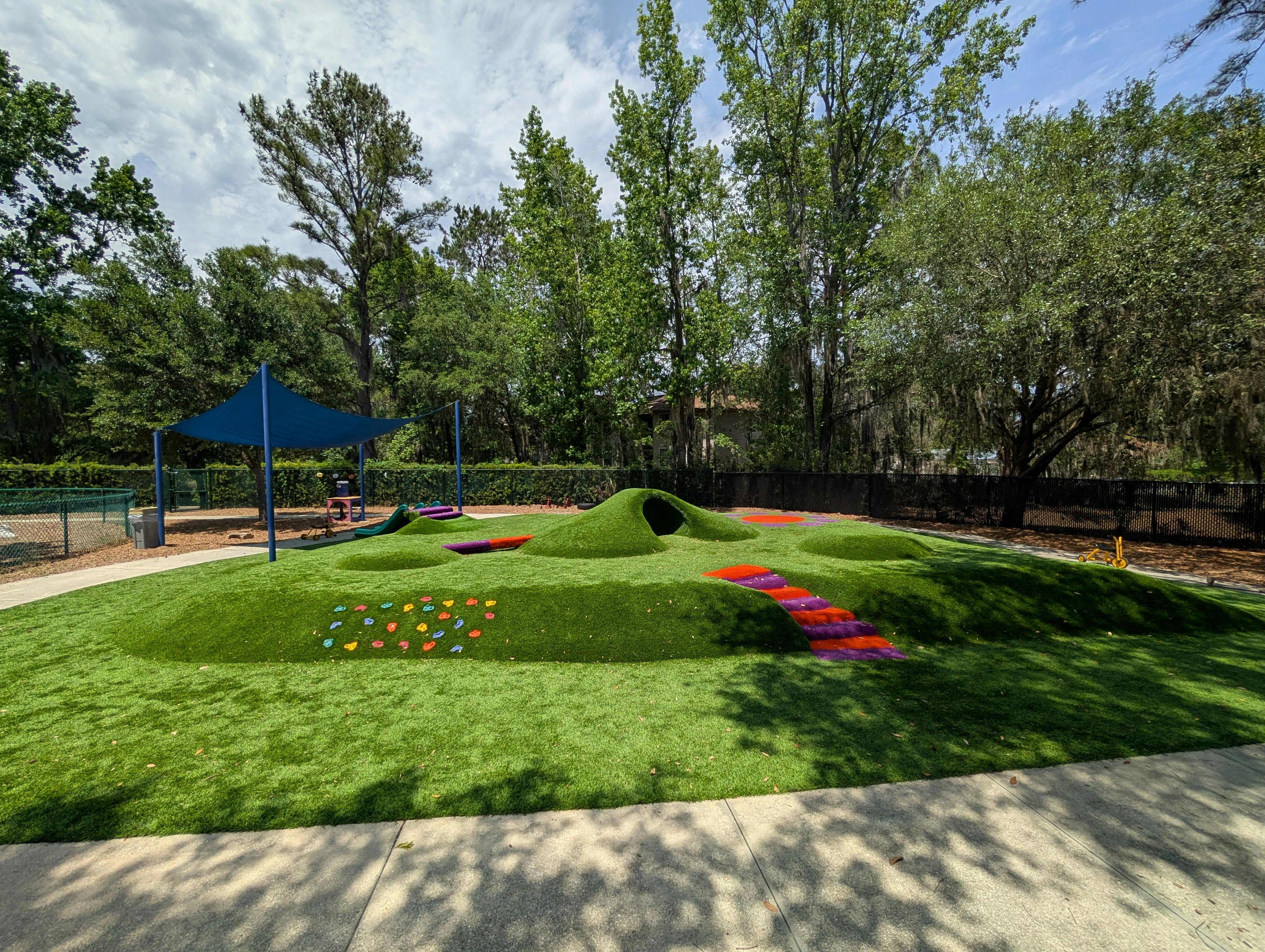 An outdoor playground with grassy mounds, a blue shade canopy, and climbing holds