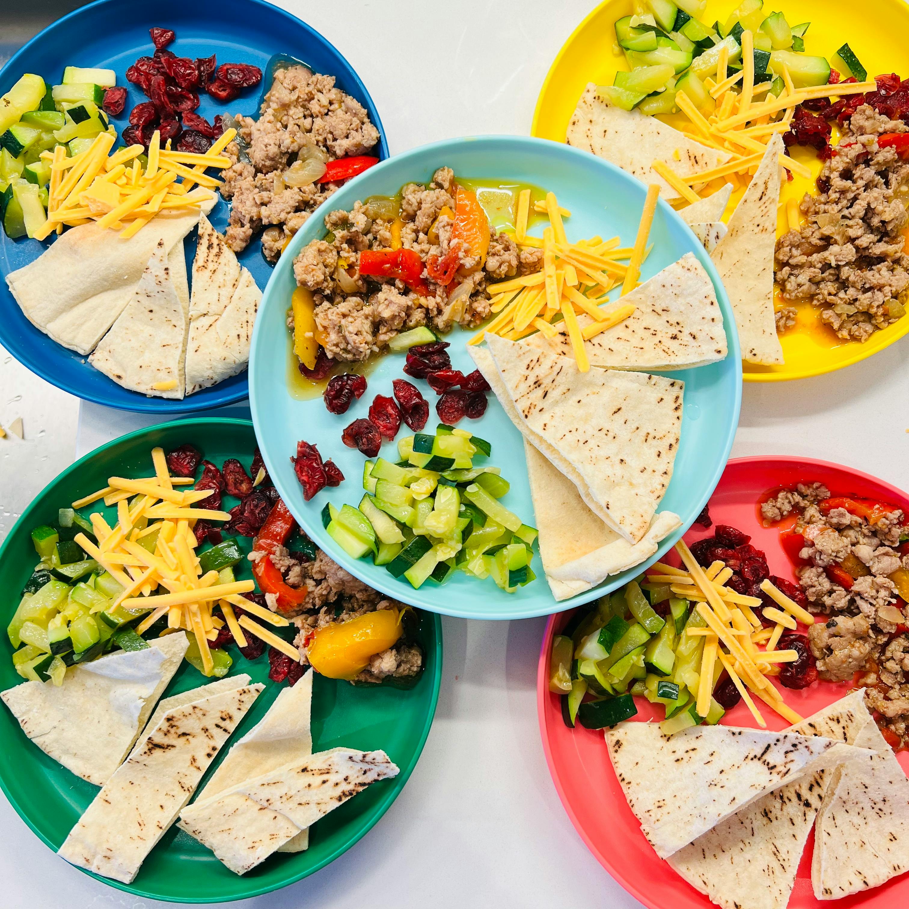 Five colorful plates of ground meat, tortillas, shredded cheese, and vegetables