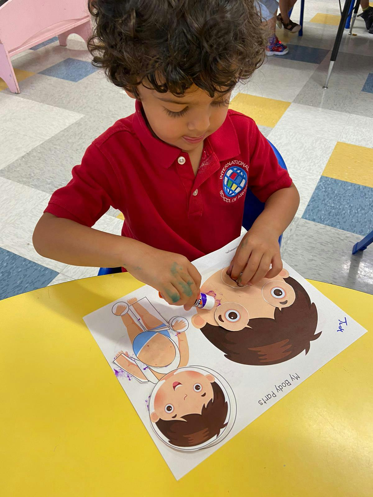 Child gluing paper cutouts onto a sheet with a drawing of a face