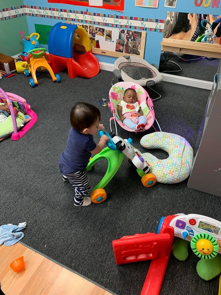 Child pushing a toy walker in a play area with another child in a bouncer