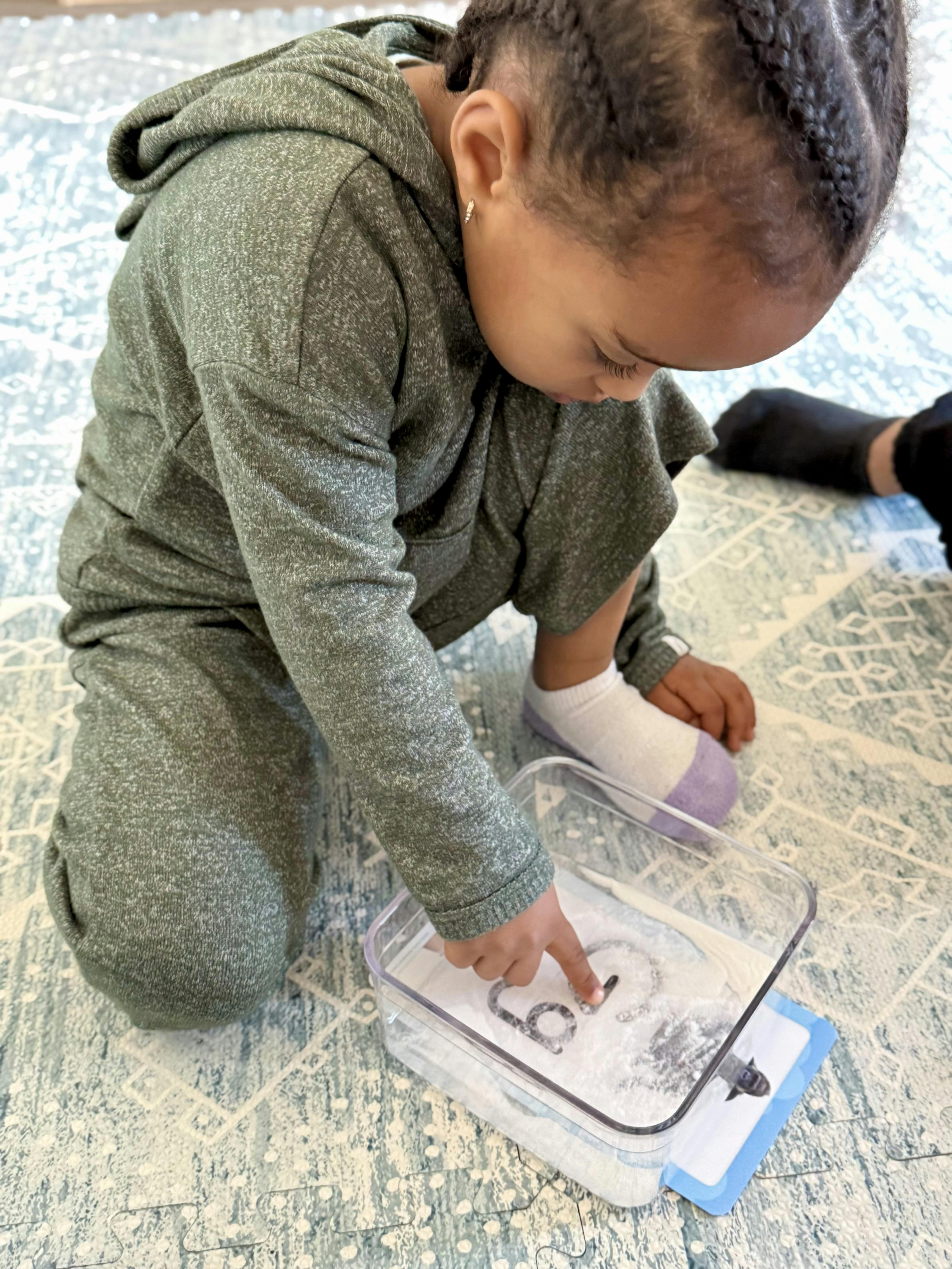 Child kneeling and tracing letters in a clear container with a finger