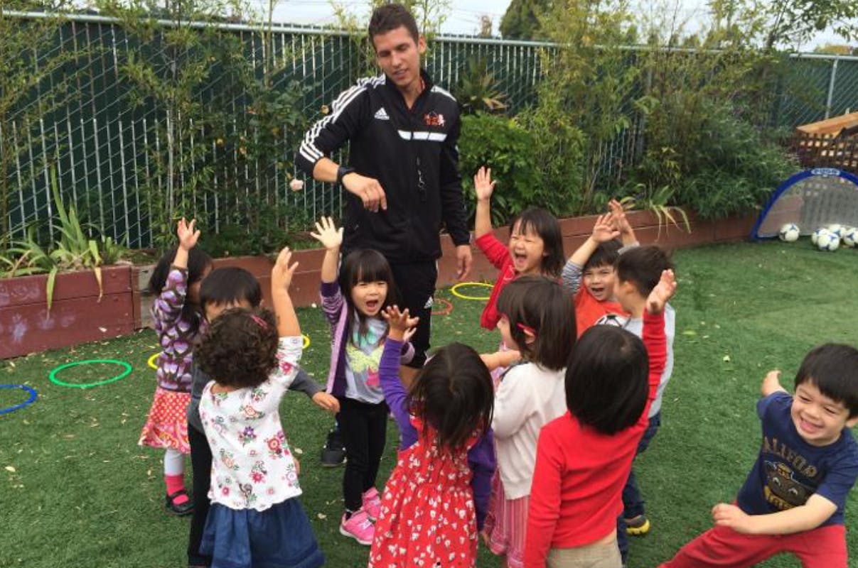 Adult and multiple children playing outdoors, many with hands raised