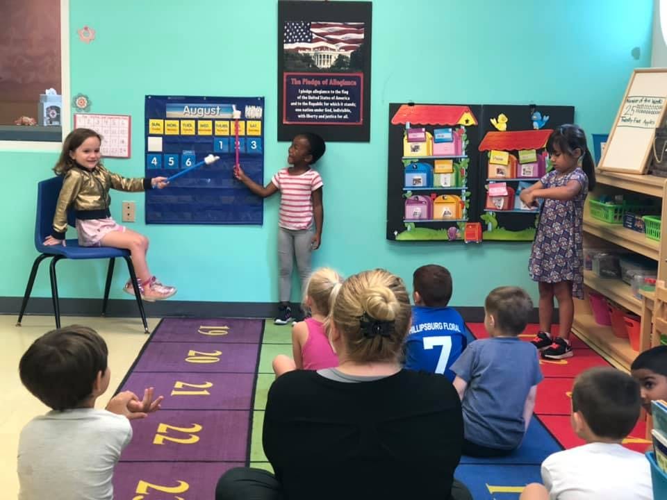 Children and an adult in a classroom, two children pointing at a wall chart