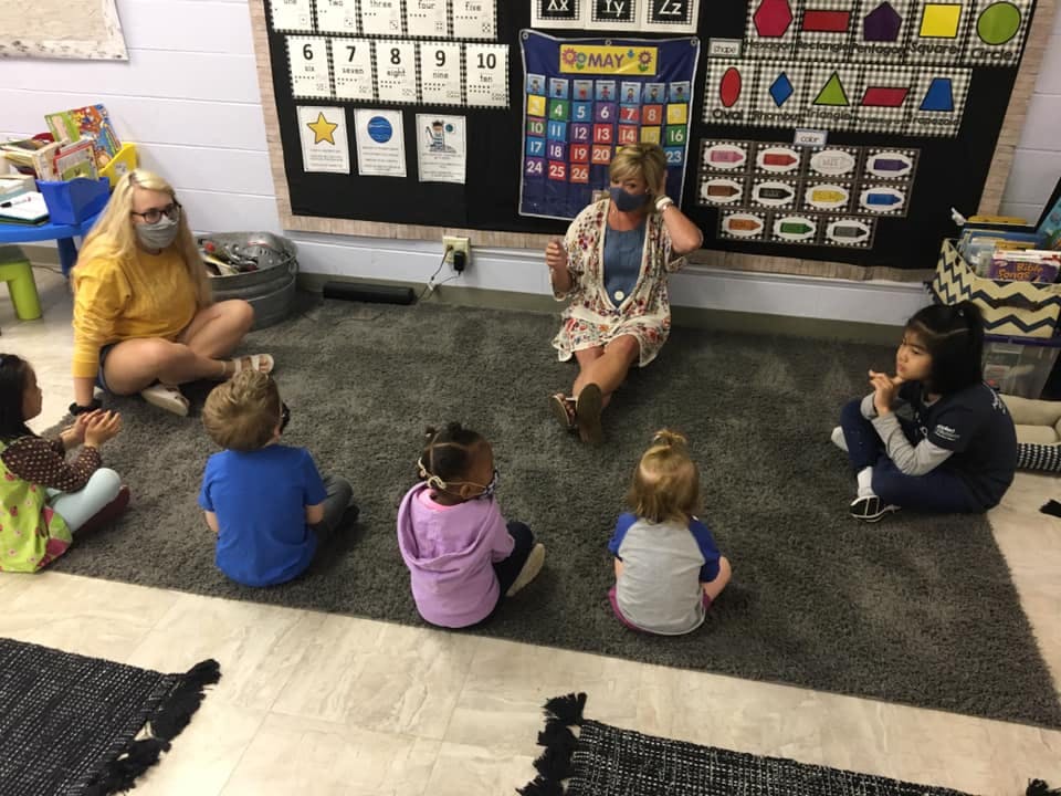 Two people sitting on a rug with several children in a classroom