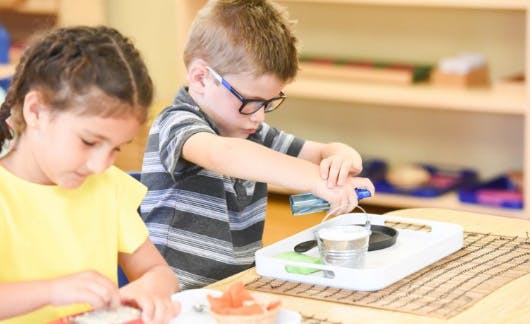 Child in glasses using a grater, another child looking down