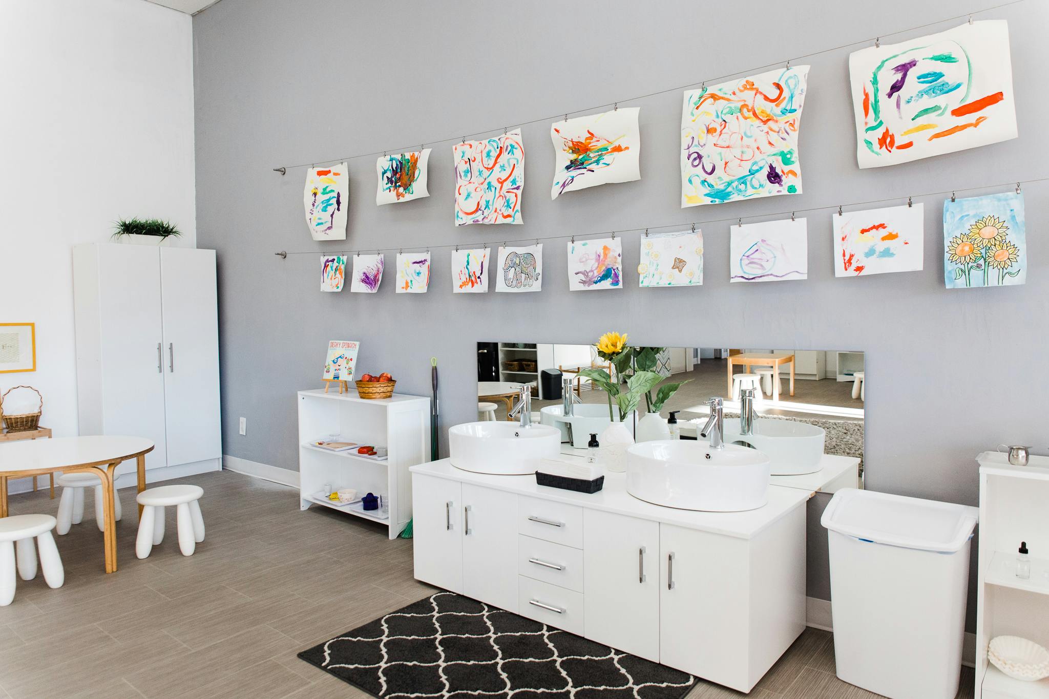 Children's artwork displayed on a wall above sinks and a small table