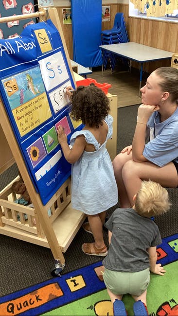 Child writing on a board with a person and another child nearby in a classroom