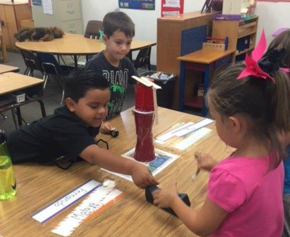 Three children playing with cups and craft sticks at a table