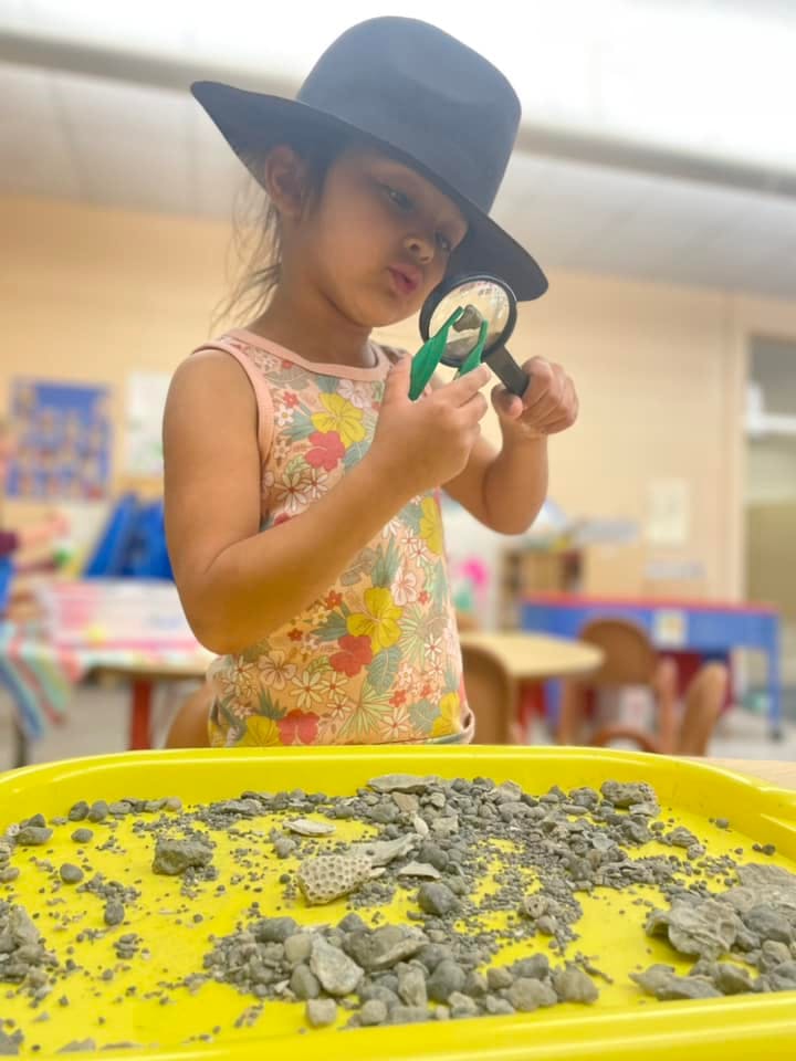 Child in a hat looking through a magnifying glass at rocks in a yellow tray