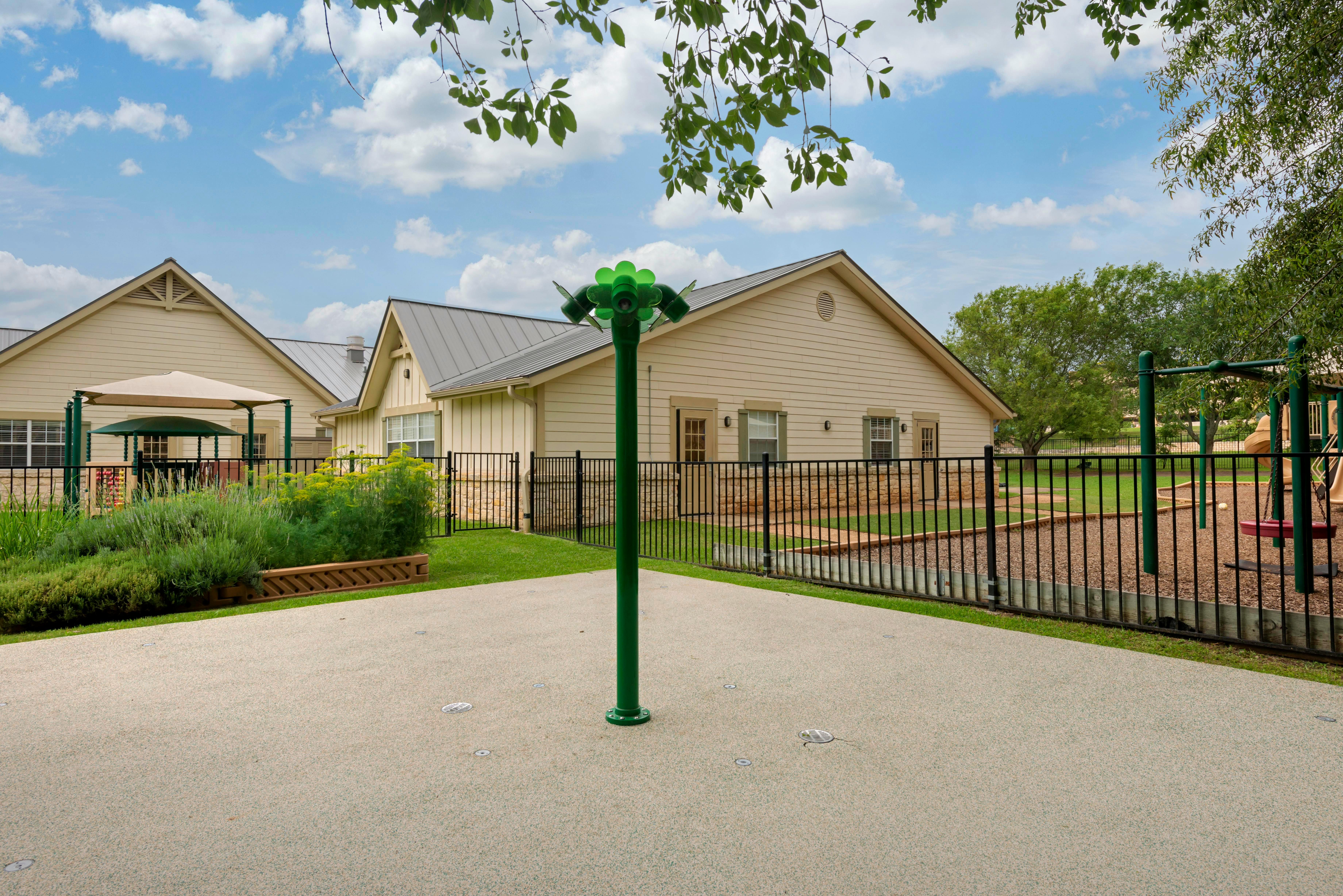A splash pad with a green flower-shaped sprinkler next to a playground with a fence