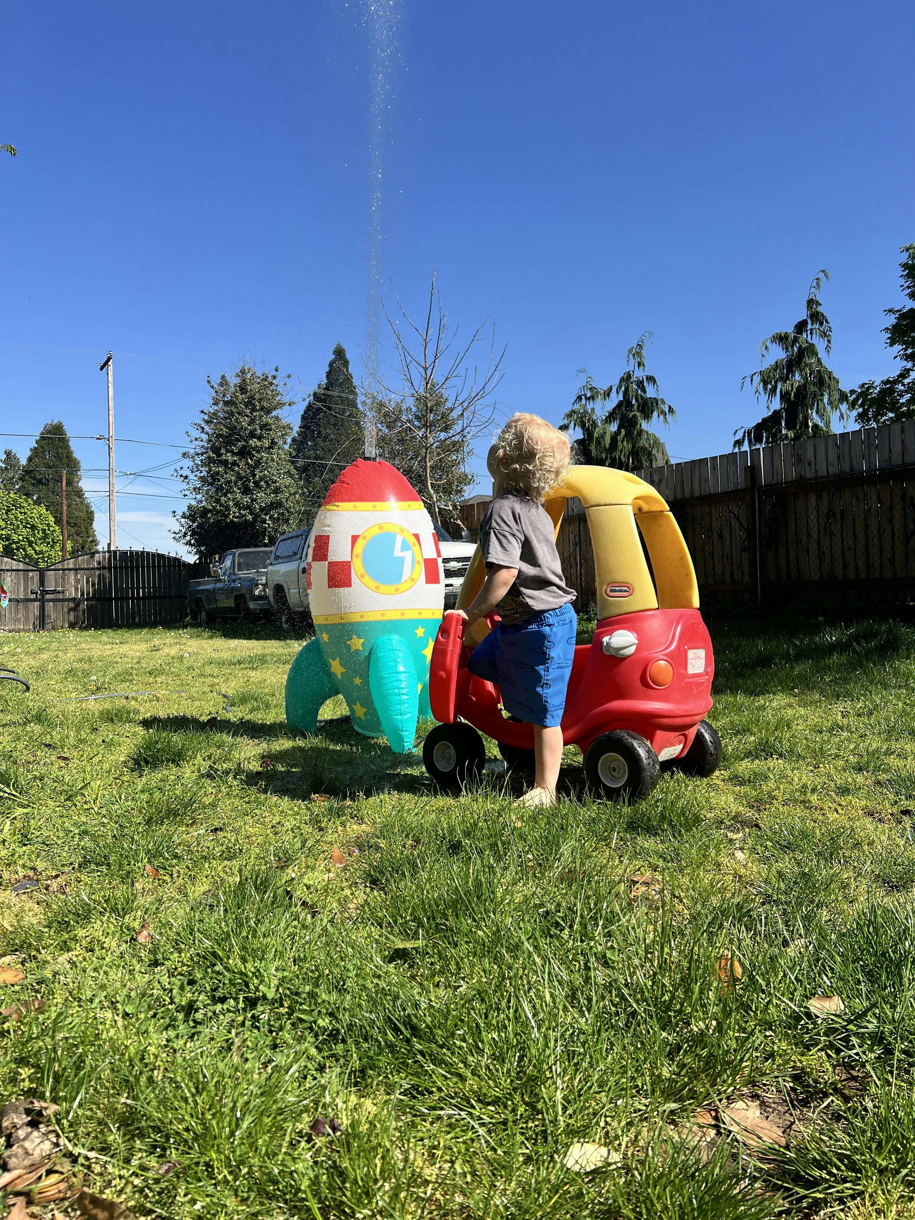 Child standing in a toy car next to a rocket-shaped lawn sprinkler
