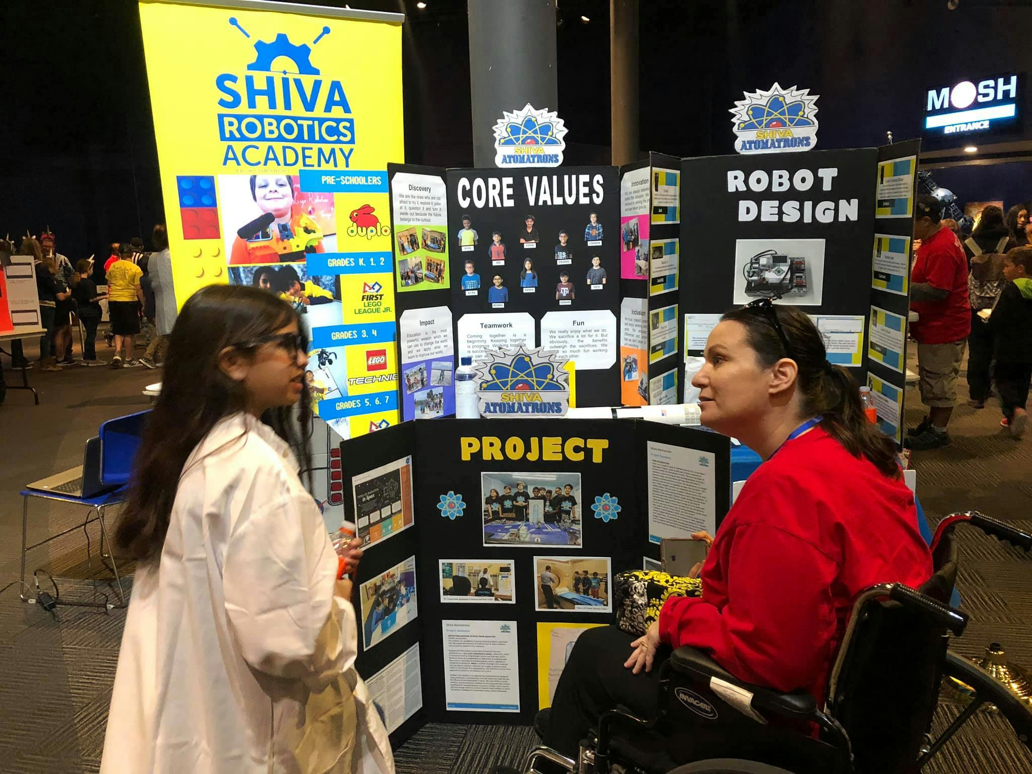 Child in lab coat talking to person in wheelchair at a project display