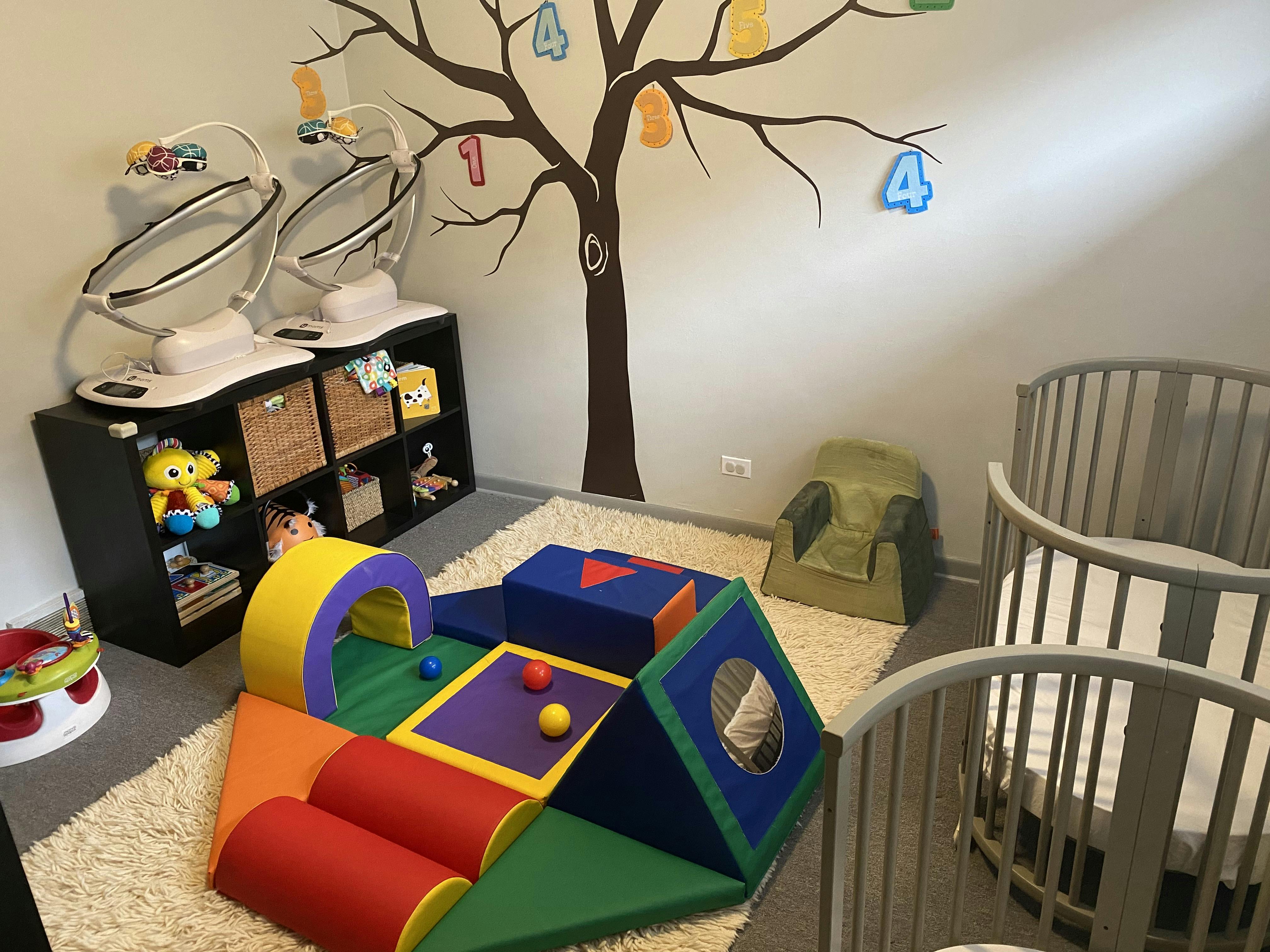 Nursery room with two cribs, soft play equipment, and a tree wall decal