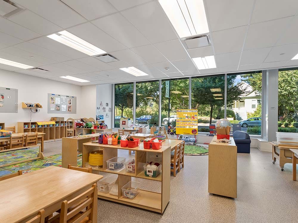 Empty classroom with small tables and chairs, shelves with toys, and large windows