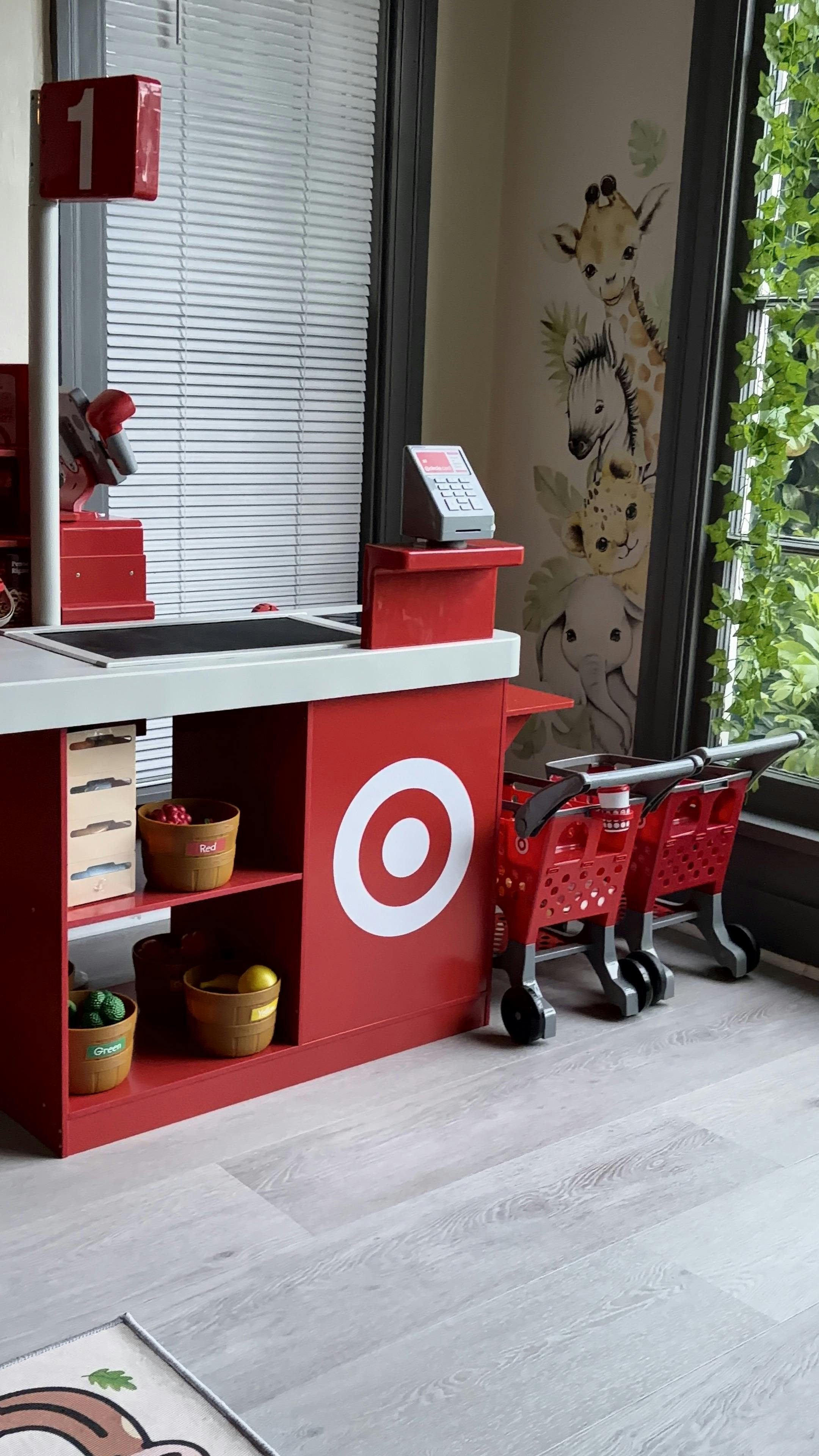 A red toy checkout counter with a Target logo, two toy shopping carts, and faux produce