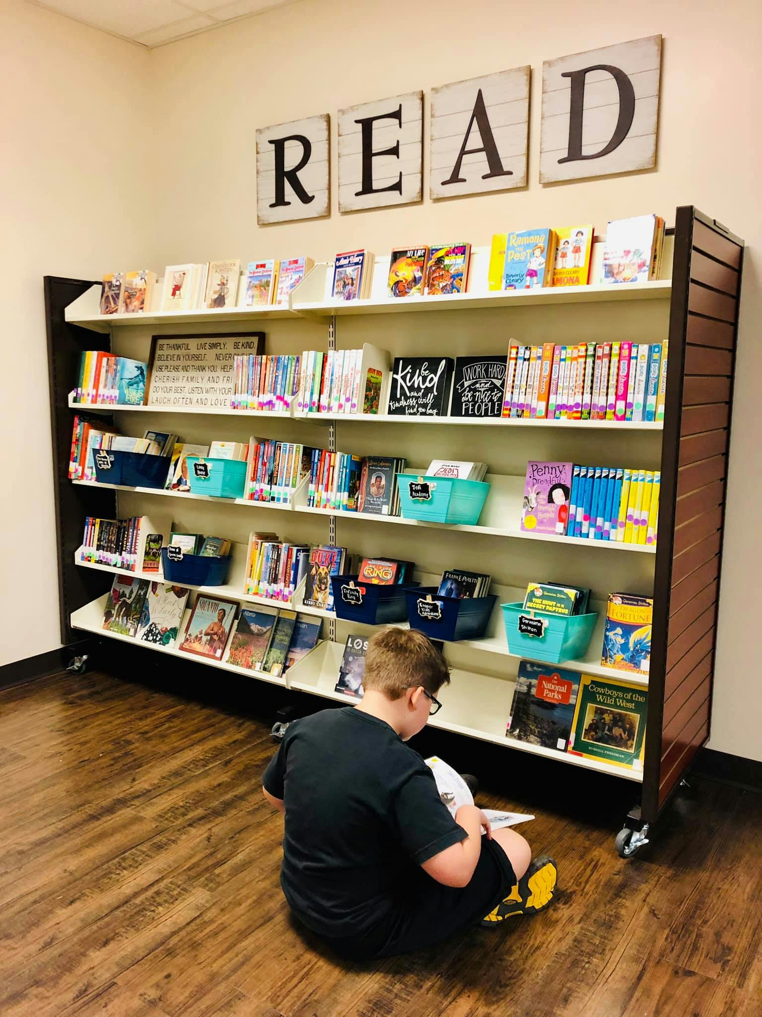 Child sitting on floor reading a book in front of a bookshelf with "READ" sign