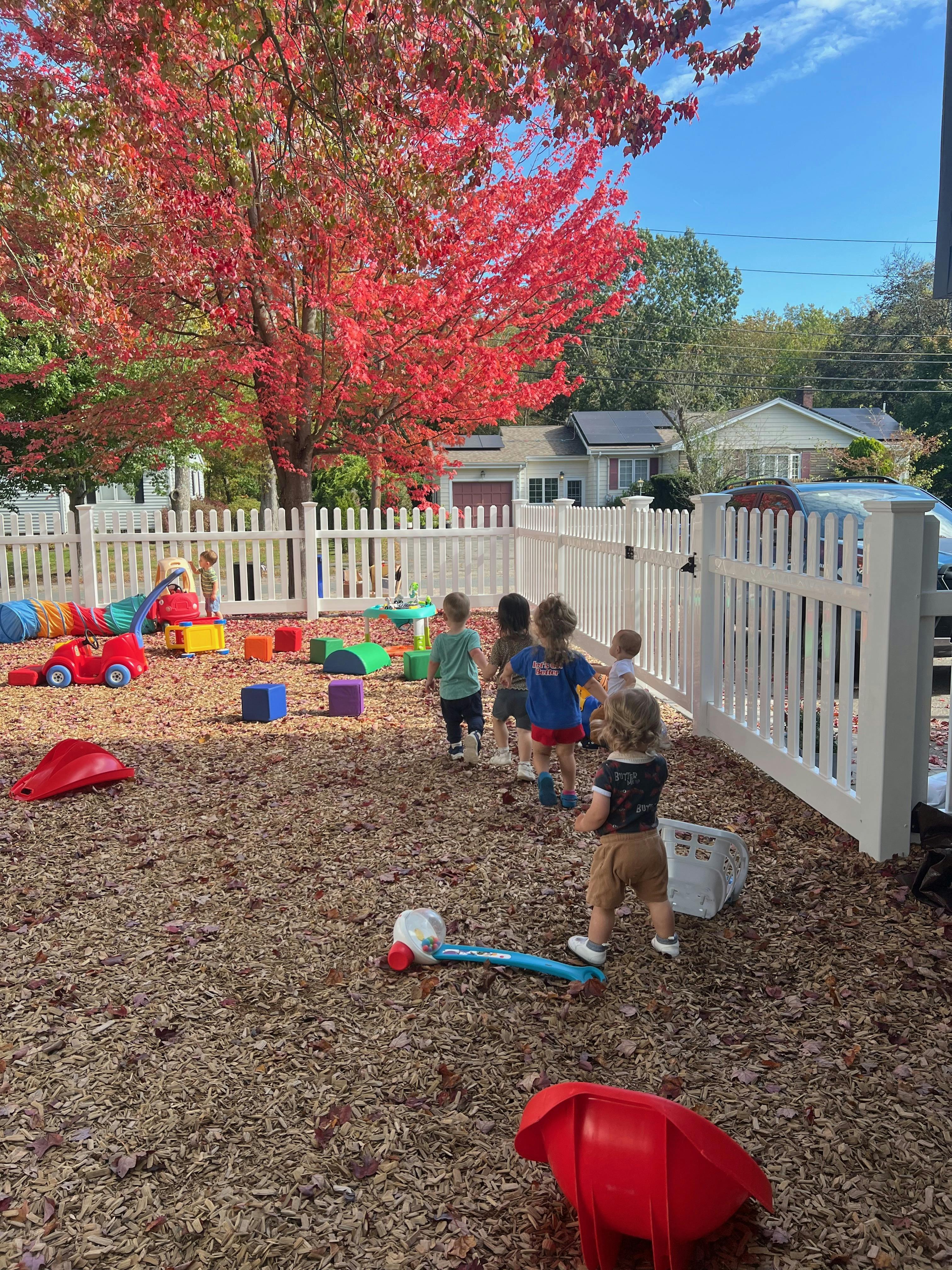Several young children playing on wood chips in an outdoor fenced-in area