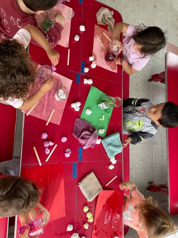 Several children doing a craft activity at a red table with jars, cotton balls, and paper
