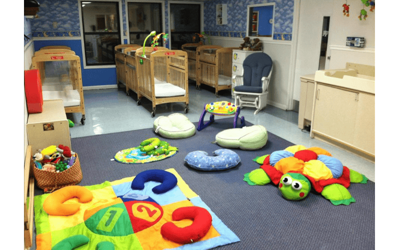 A colorful baby room with multiple cribs, a playmat, cushions, and toys