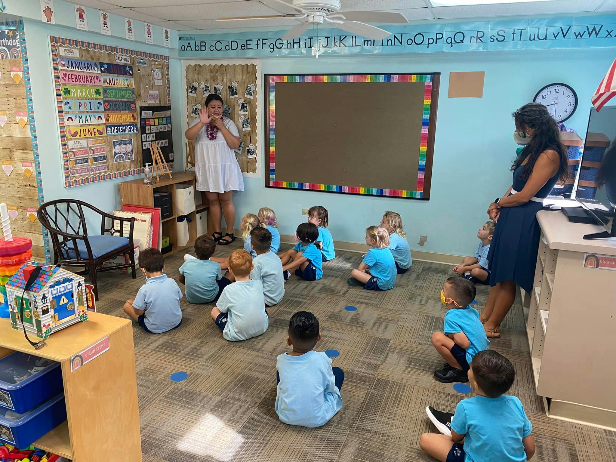 Two people standing in a classroom with children sitting on the floor