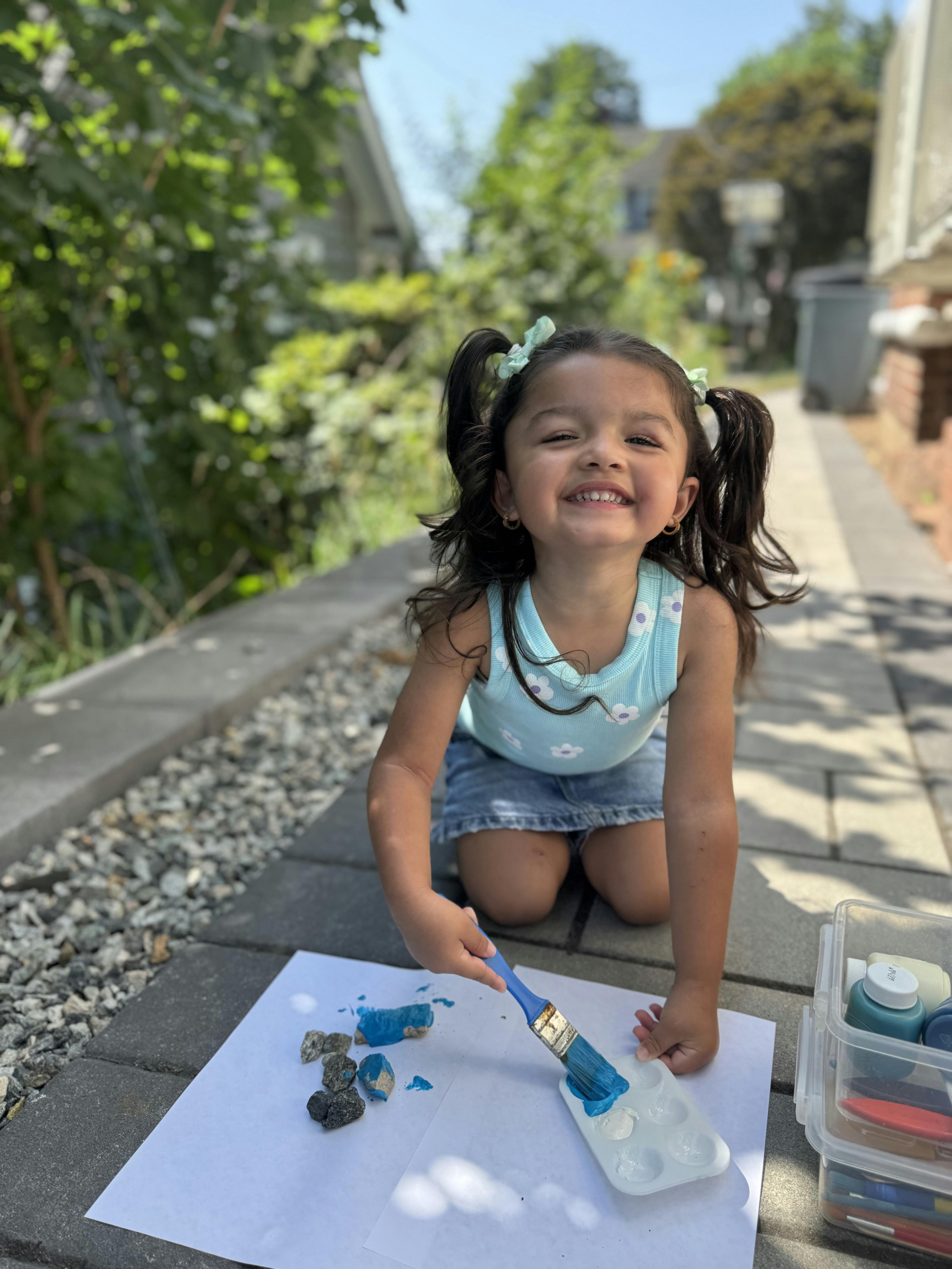 Child kneeling and painting rocks blue on a white paper outdoors