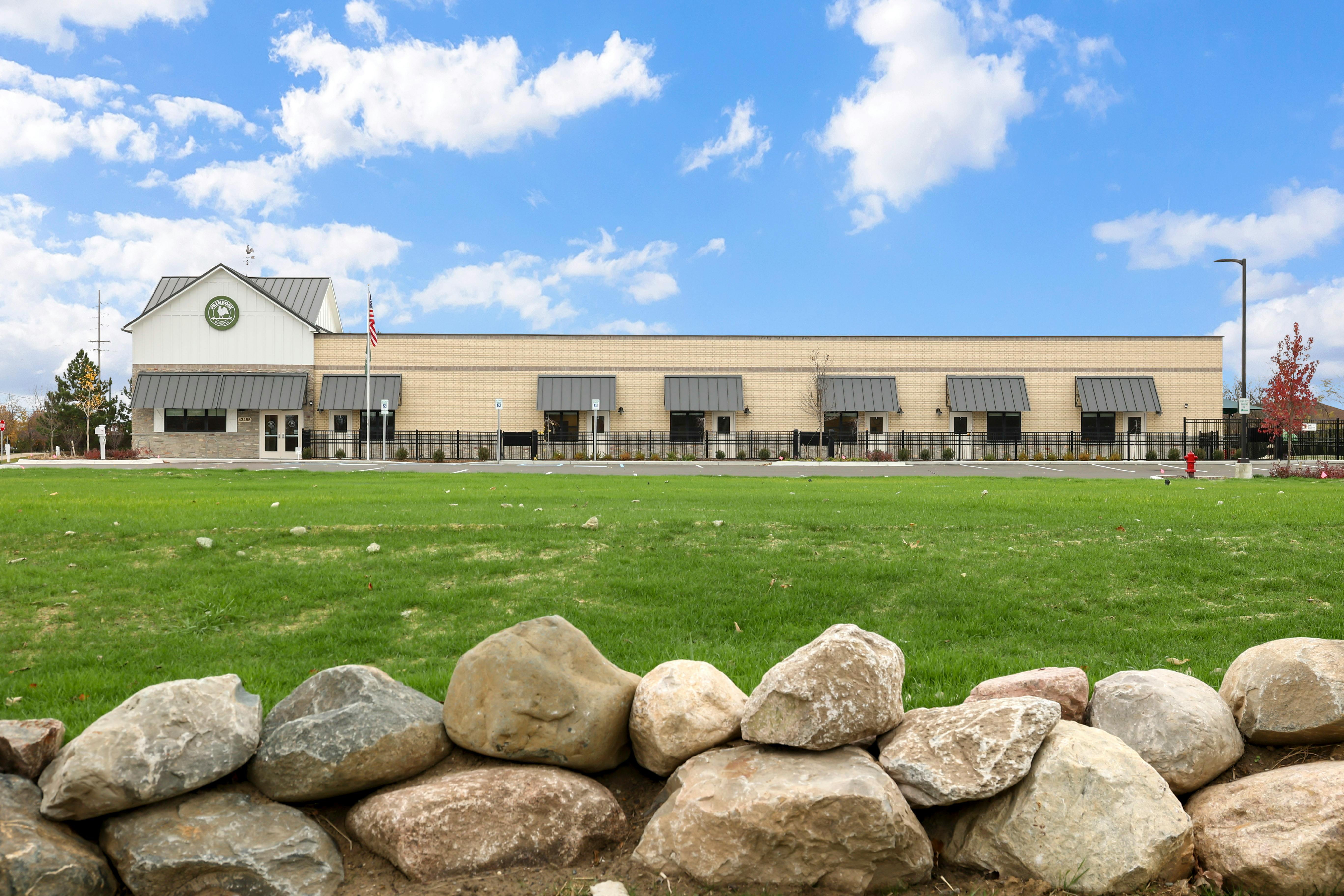 Tan brick building with a gray roof and awnings, with green grass and rocks in front