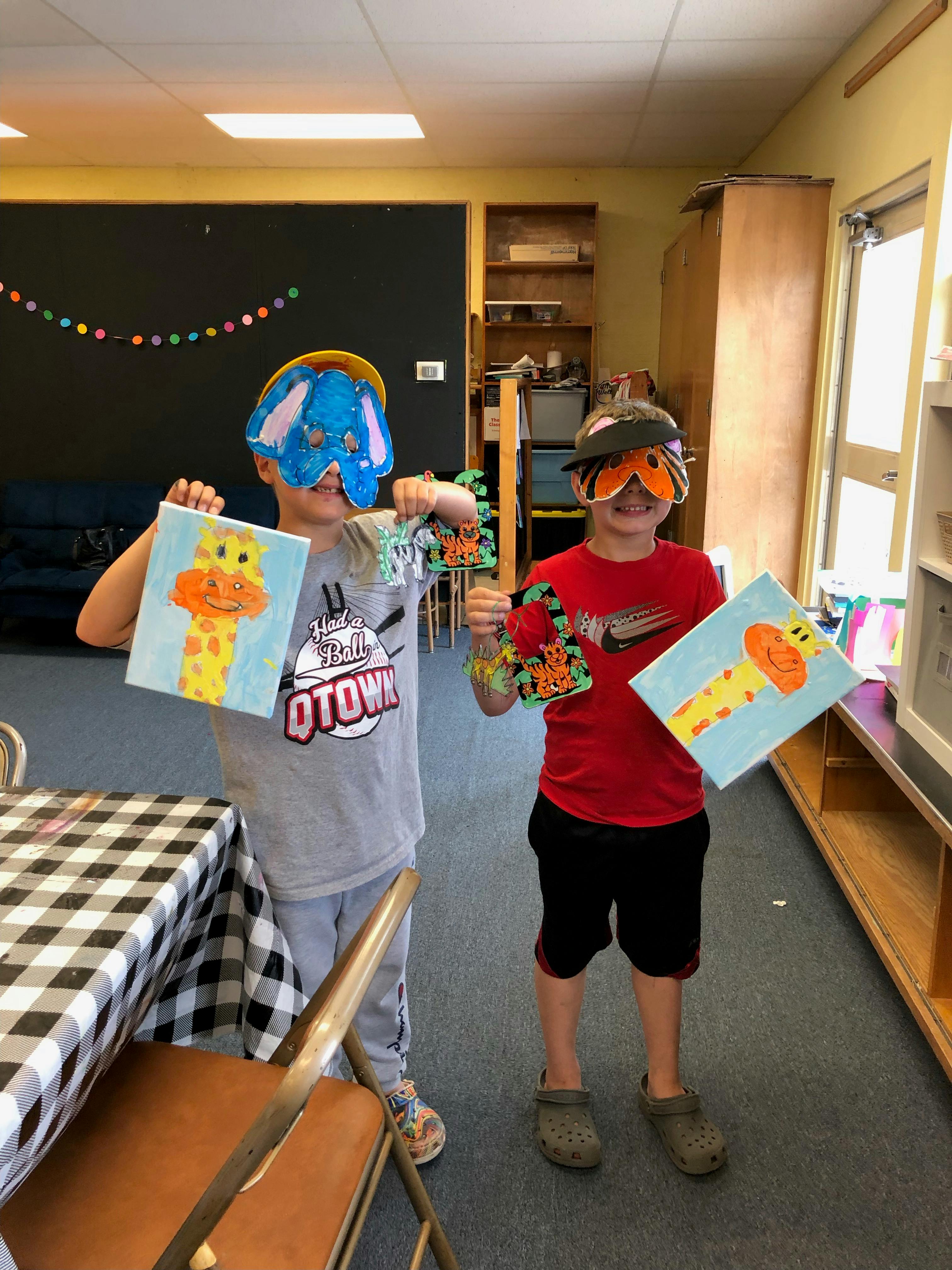 Two children wearing masks and holding up paintings and crafts