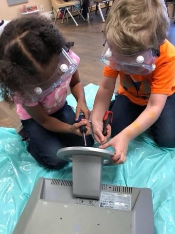 Two children in safety goggles using screwdrivers on a computer monitor