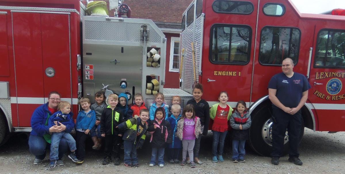 Two adults and multiple children posing in front of two fire trucks