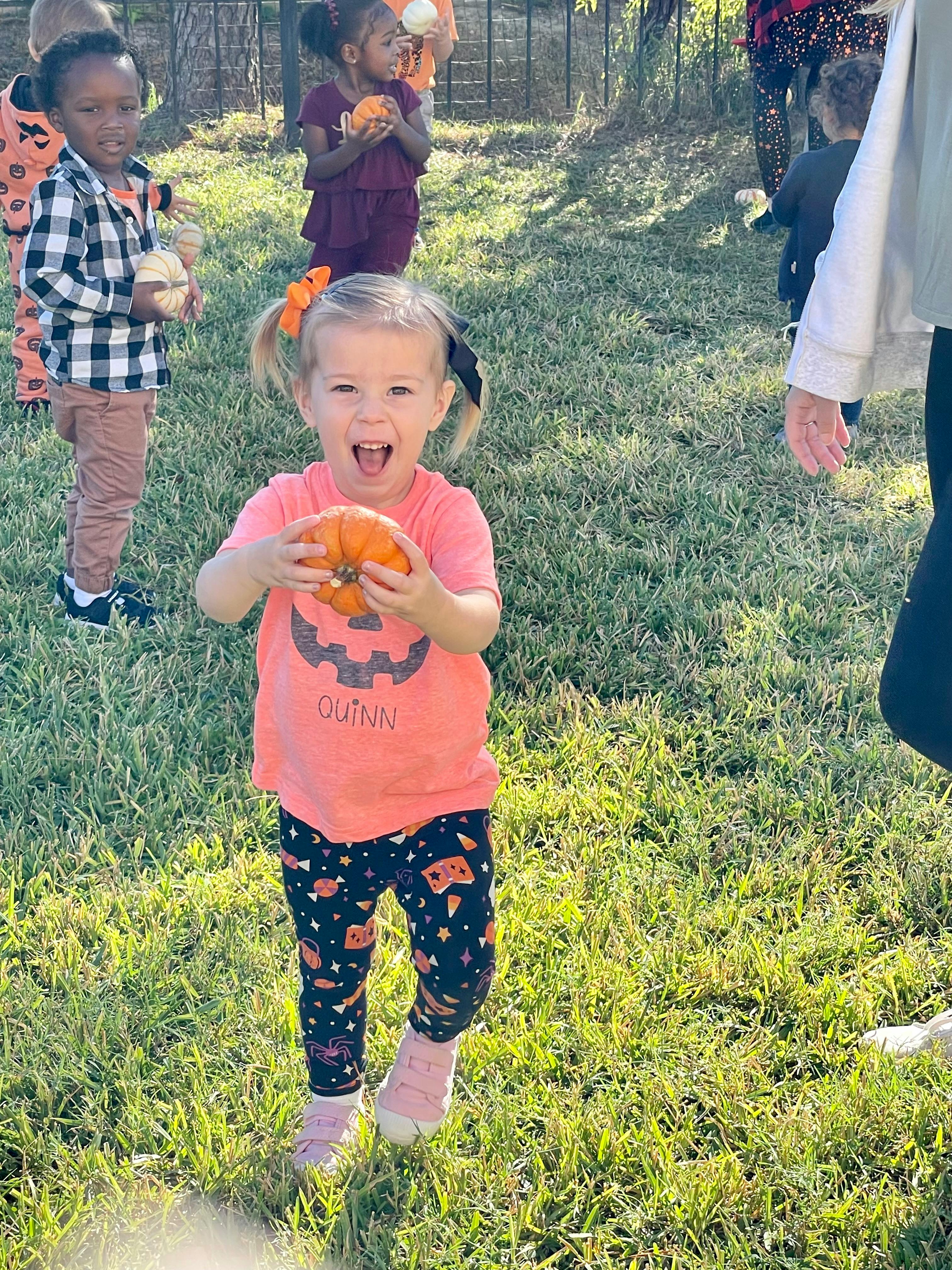 Child holding a small pumpkin outdoors with other children in the background