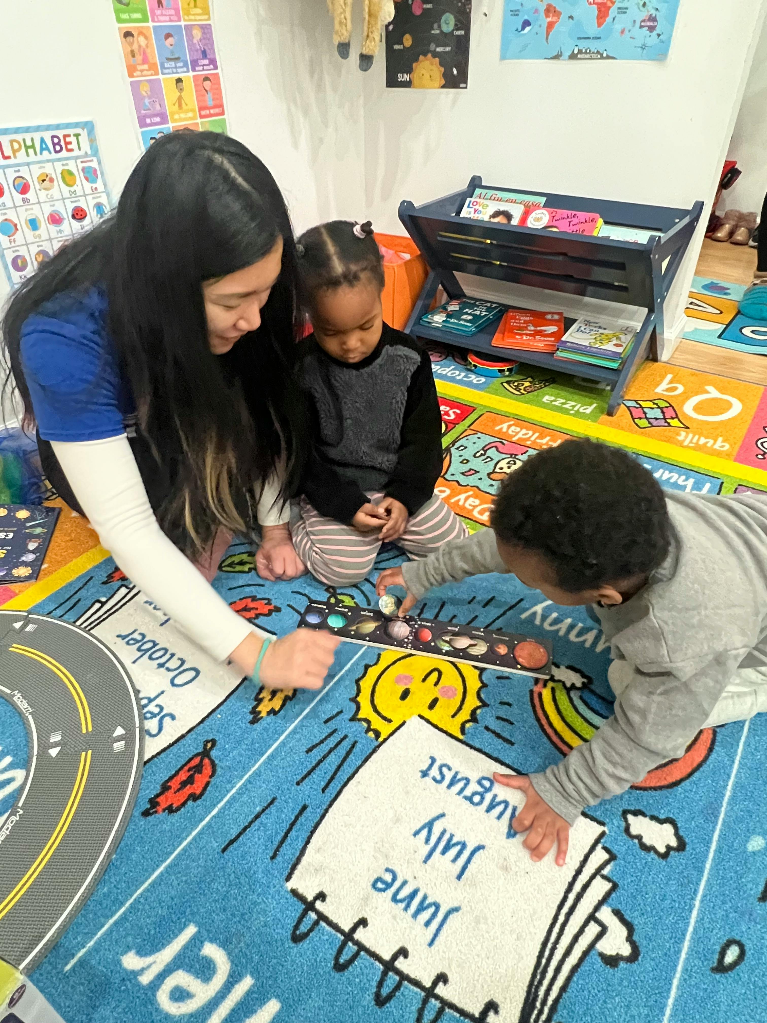 Person and two children playing on a colorful rug in a classroom