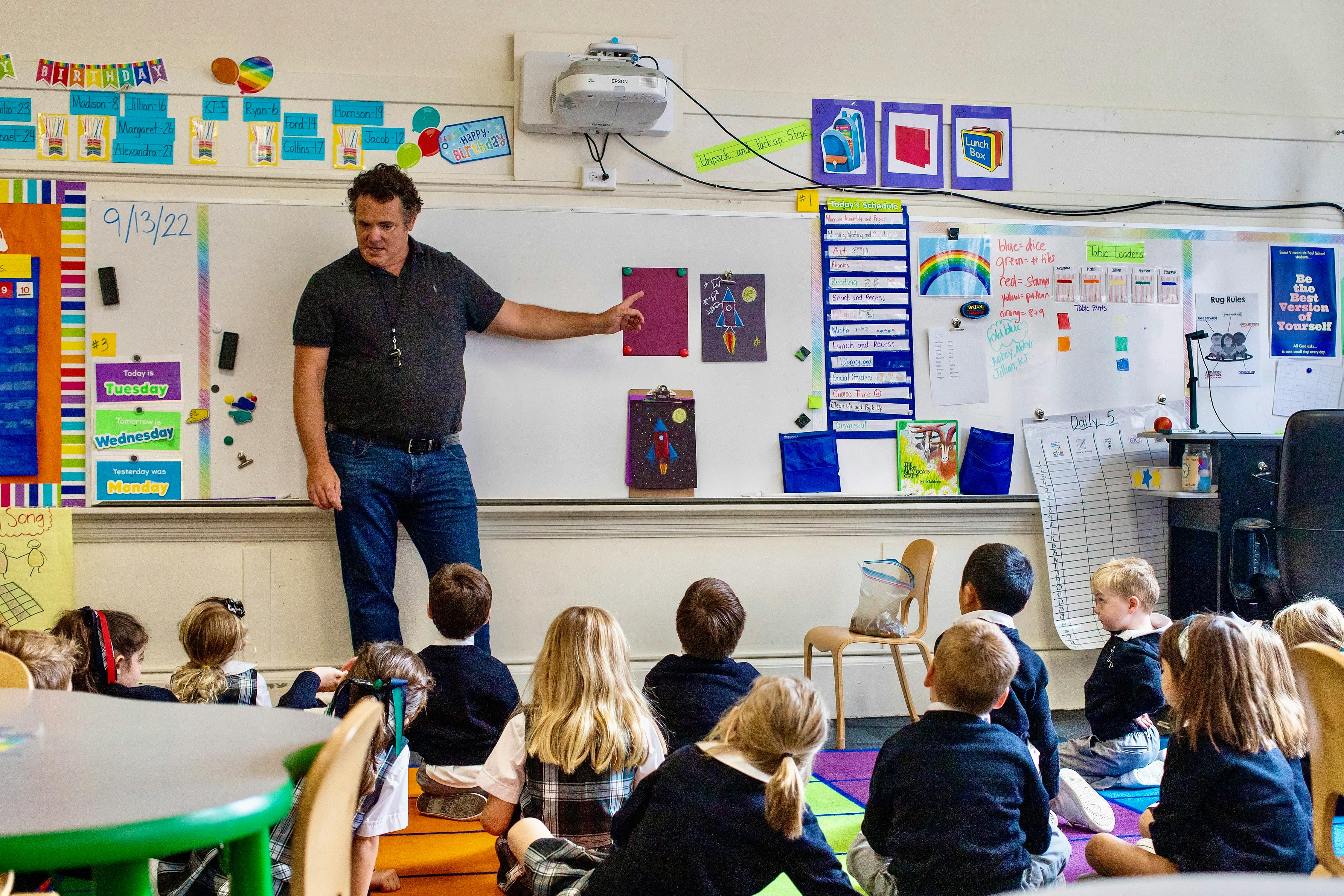 Adult male teacher pointing at a whiteboard with children sitting on the floor in front