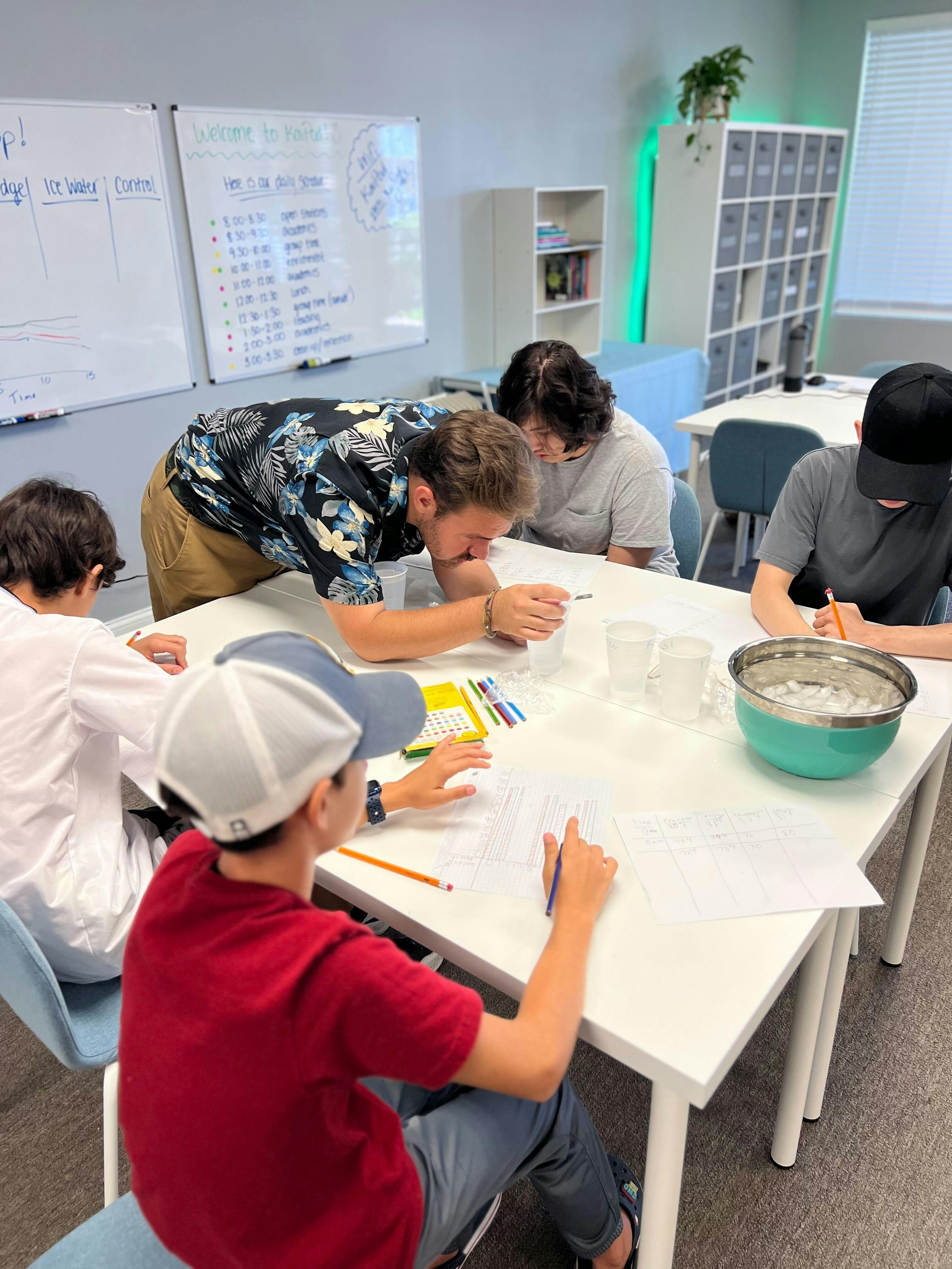People gathered around a table in a classroom, some writing and observing items onlooking