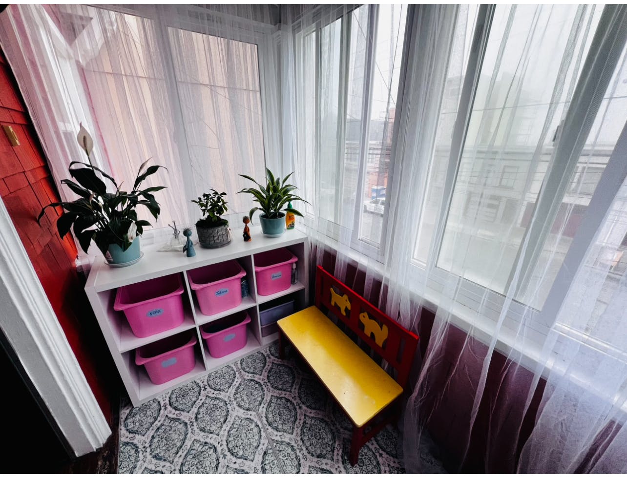 Sunroom with white shelving unit, pink bins, plants, and a yellow bench