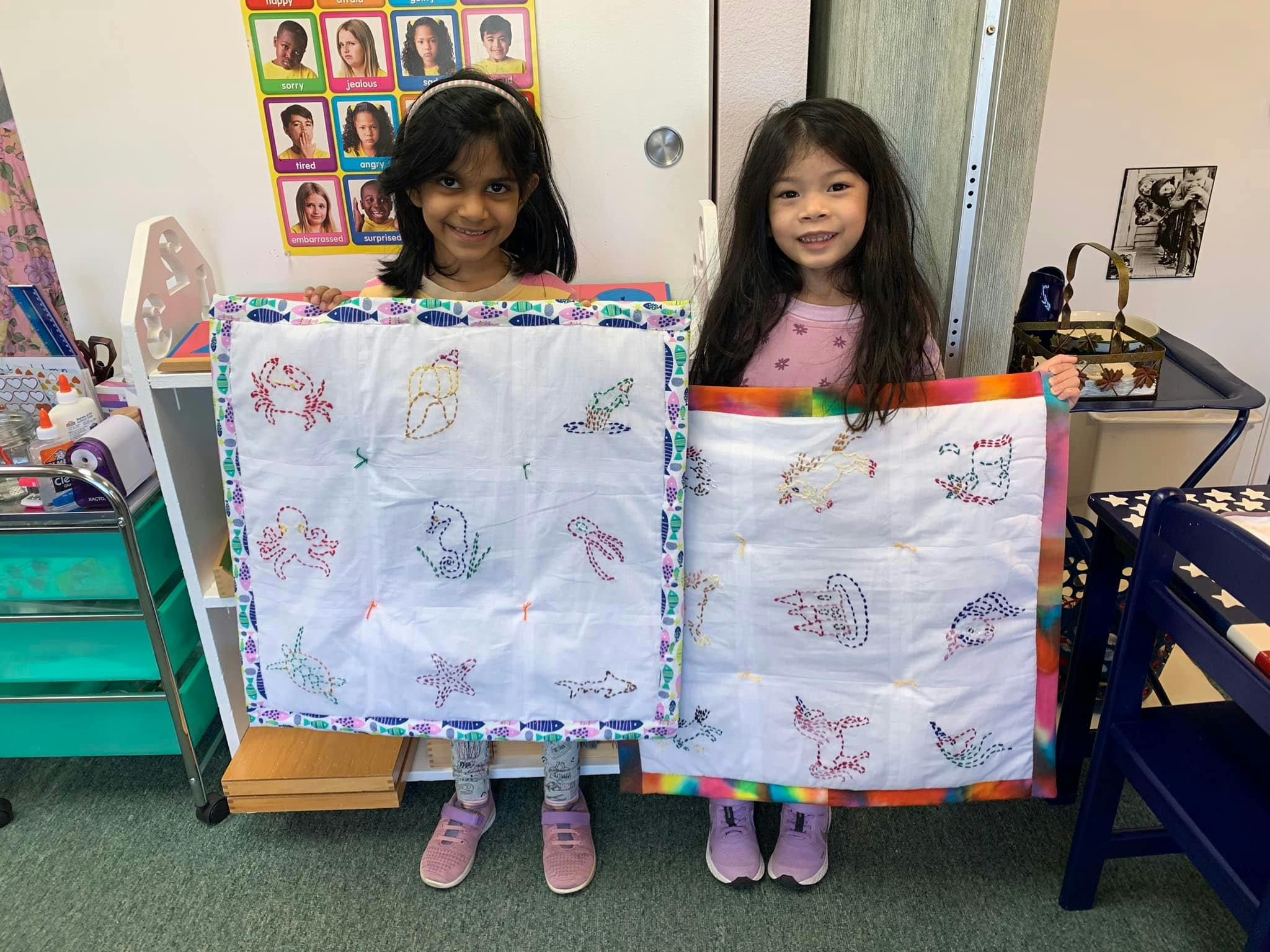 Two children holding up square fabric art pieces with drawings