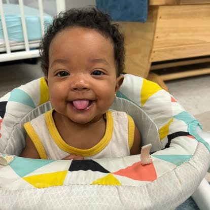 A young child sitting in an activity seat with tongue out