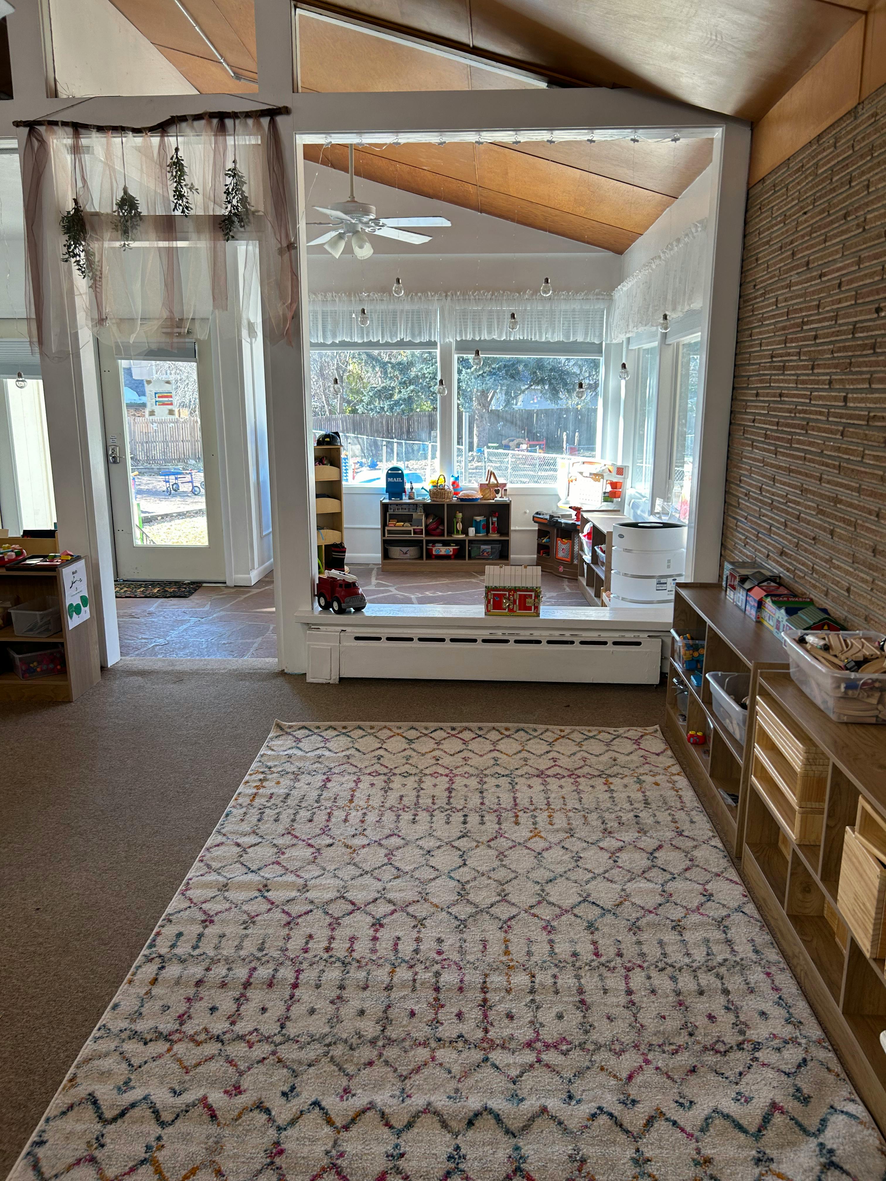 A room with a patterned rug and shelves, leading to a sunroom with large windows