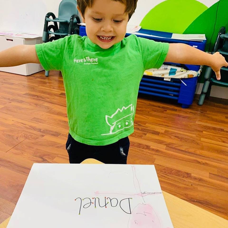 Child in a green shirt standing over a paper with the name Daniel written on it