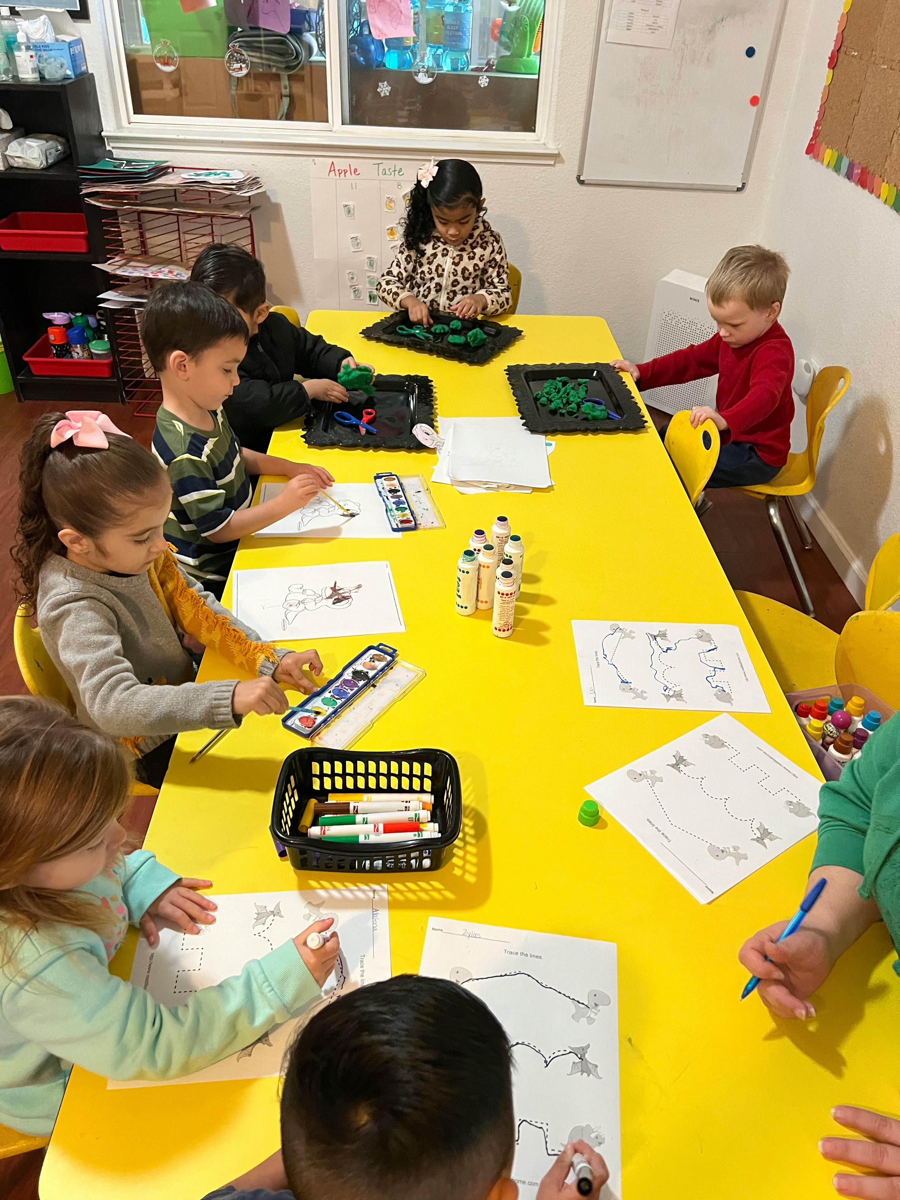 Children sitting at a yellow table, drawing and playing with small objects