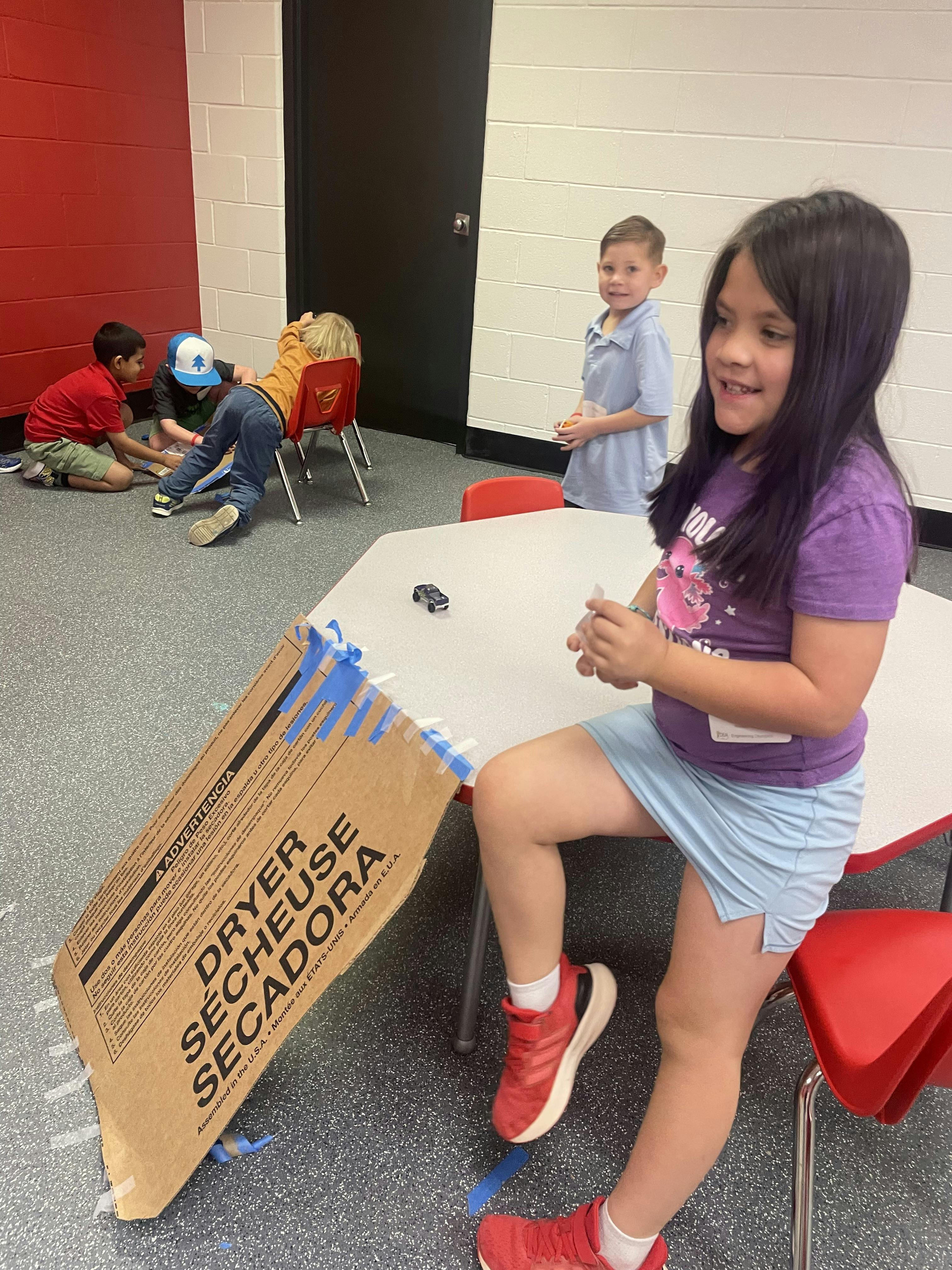 Children playing with a cardboard ramp and toy car at a table