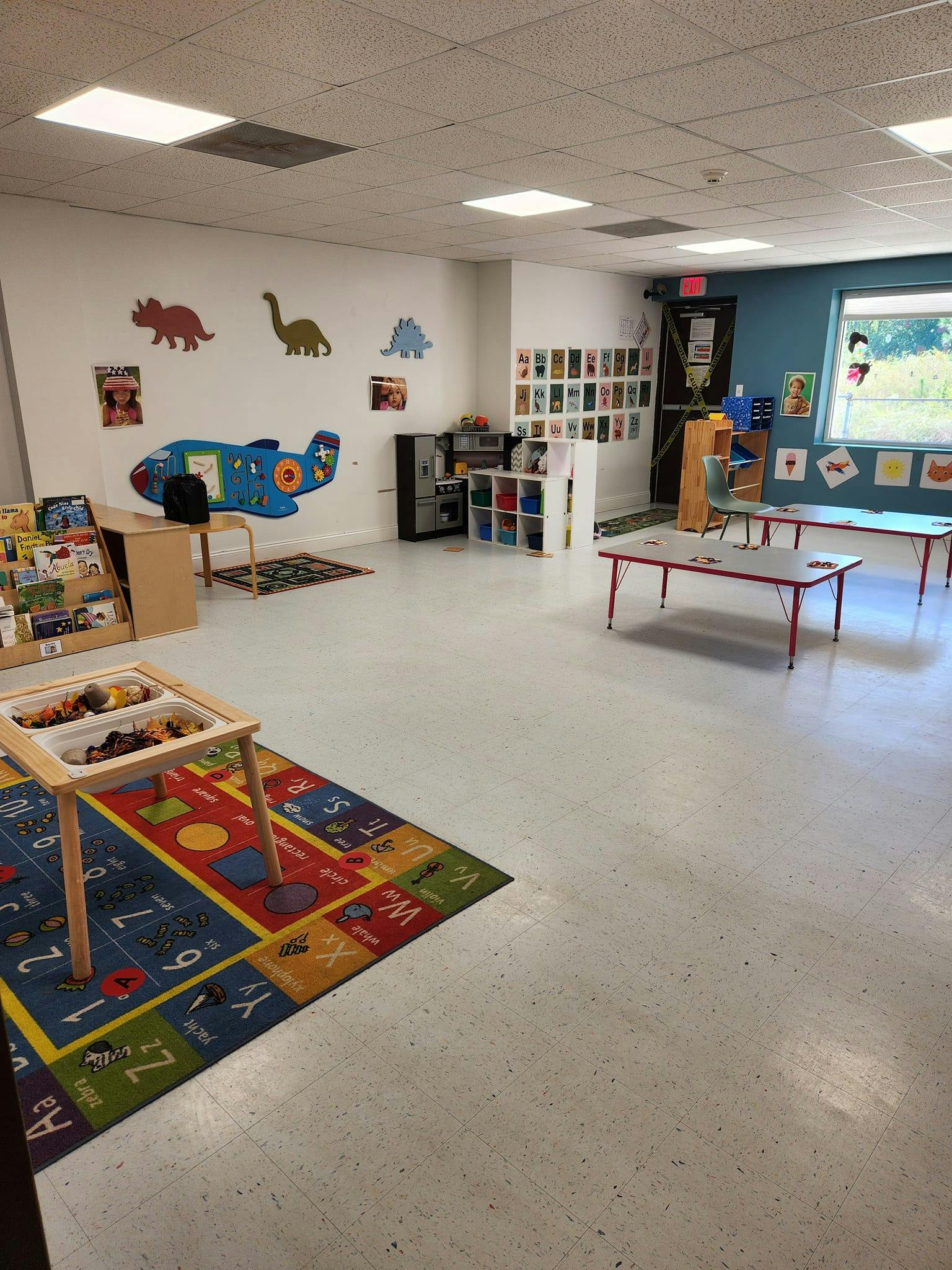 A colorful classroom with play tables, rugs, and dinosaur cutouts on the wall