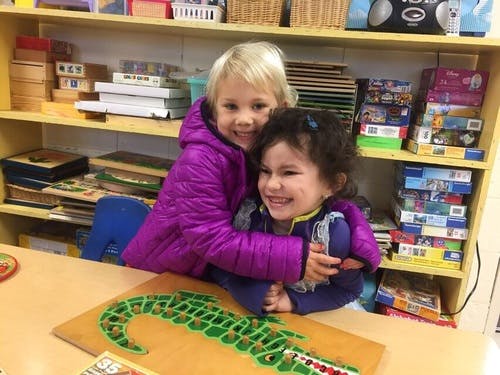 Two children embracing in a classroom with a puzzle on a table