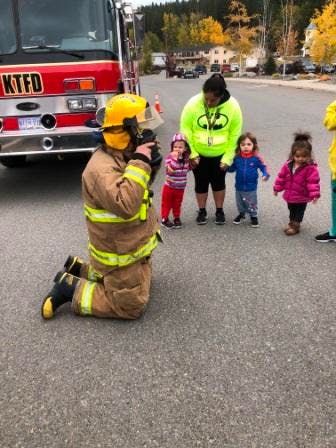 Firefighter kneeling in front of children and an adult near a fire truck