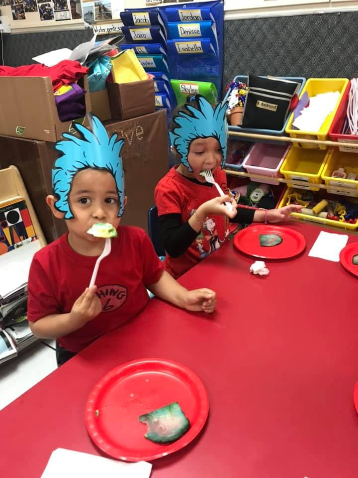 Two children wearing blue paper hats eating at a red table